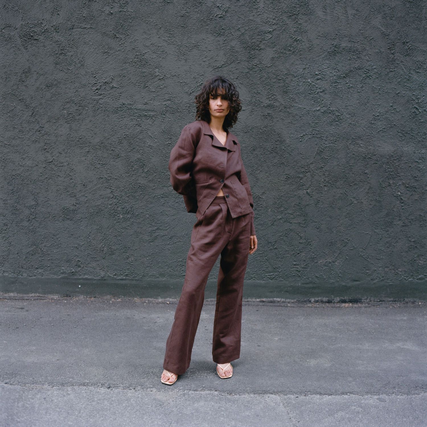 A model wears a brown linen suit from Waves of Hydra on a sidewalk in front of a grey wall.