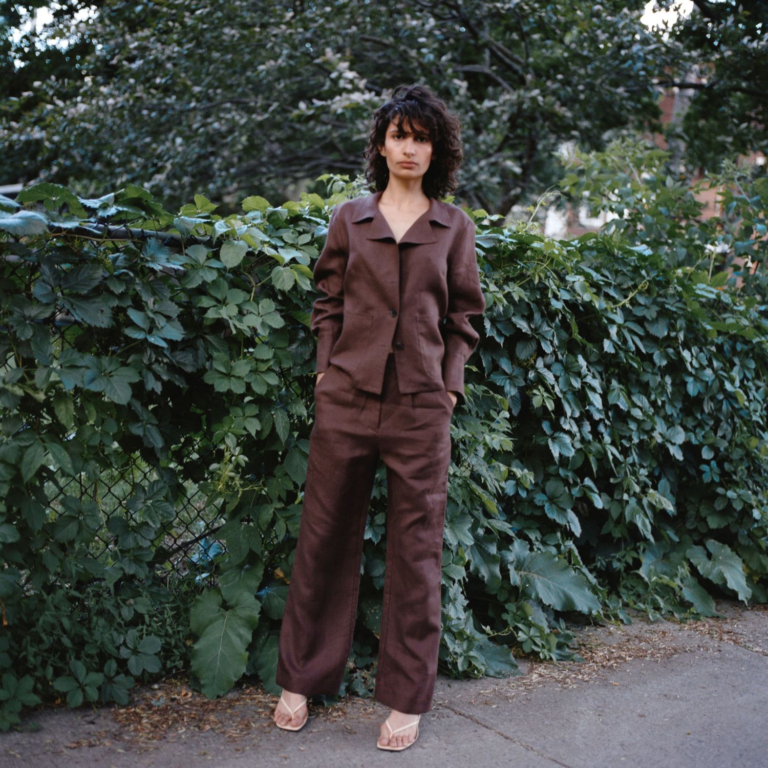 A model wears a brown linen suit from Waves of Hydra on the sidewalk surrounded by green vines and lush trees.
