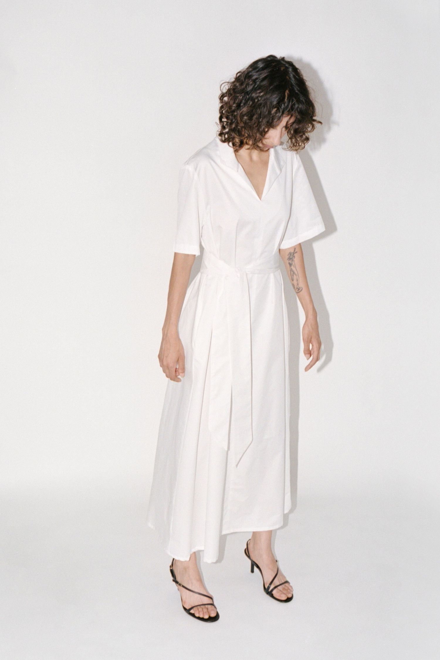 A wavy-haired brunette model wears a minimal long white cotton dress with belt from Waves of Hydra.