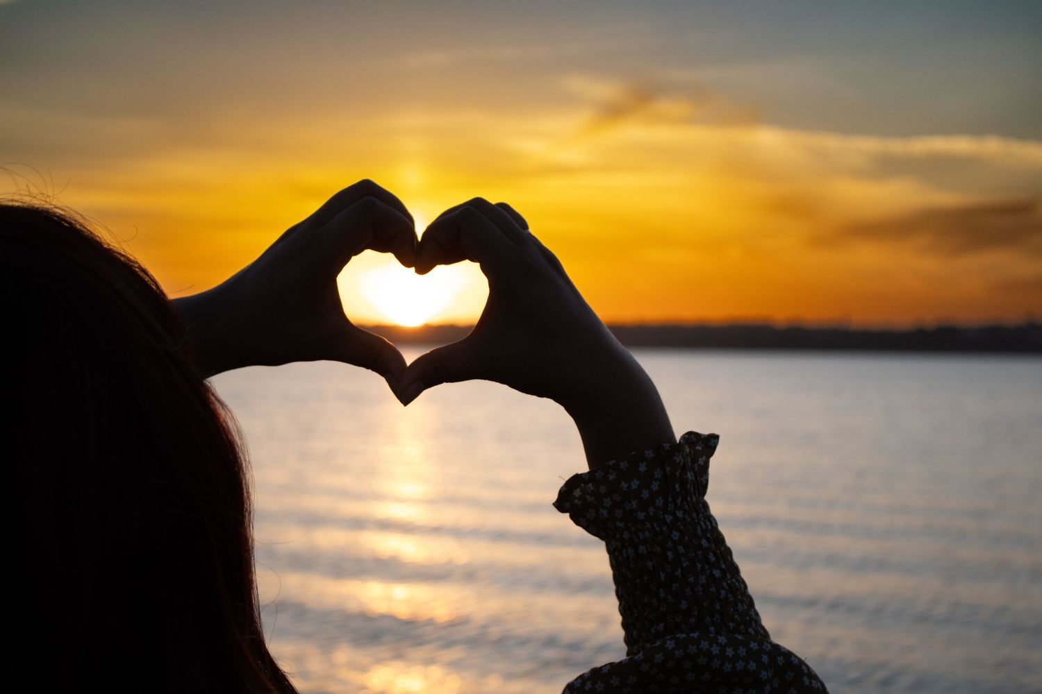 Hands forming a heart around the setting sun over the water