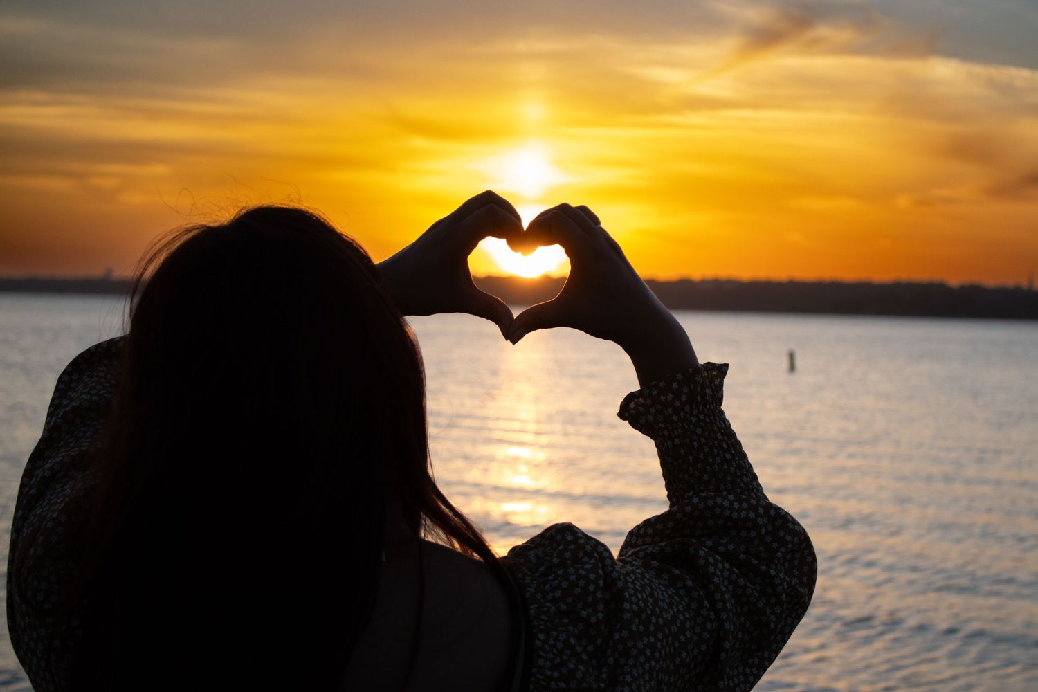 Woman making a heart with her hands during a golden sunset over the water