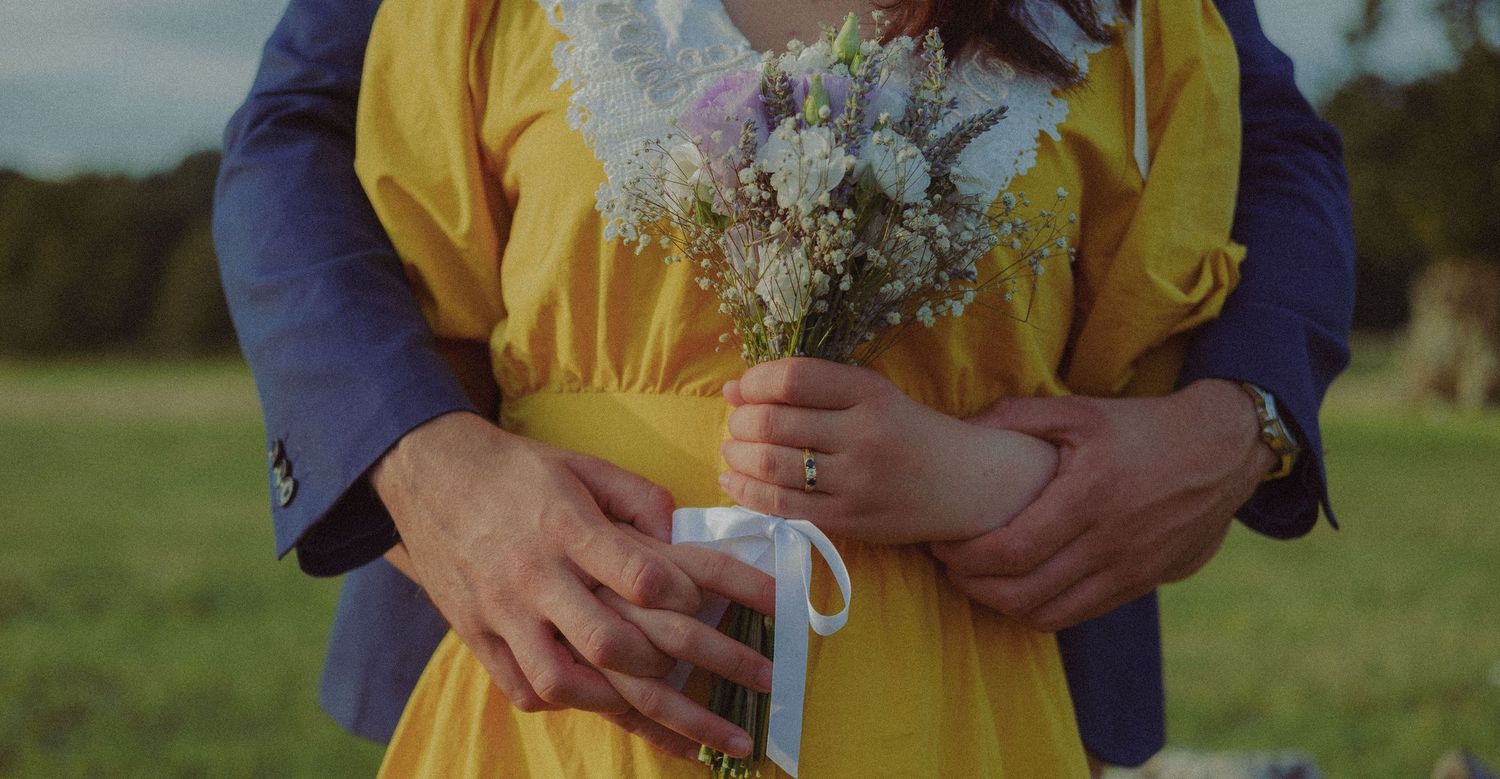 Intimate wedding photograph of a newlywed couple embracing with a bouquet of flowers in a countryside setting.