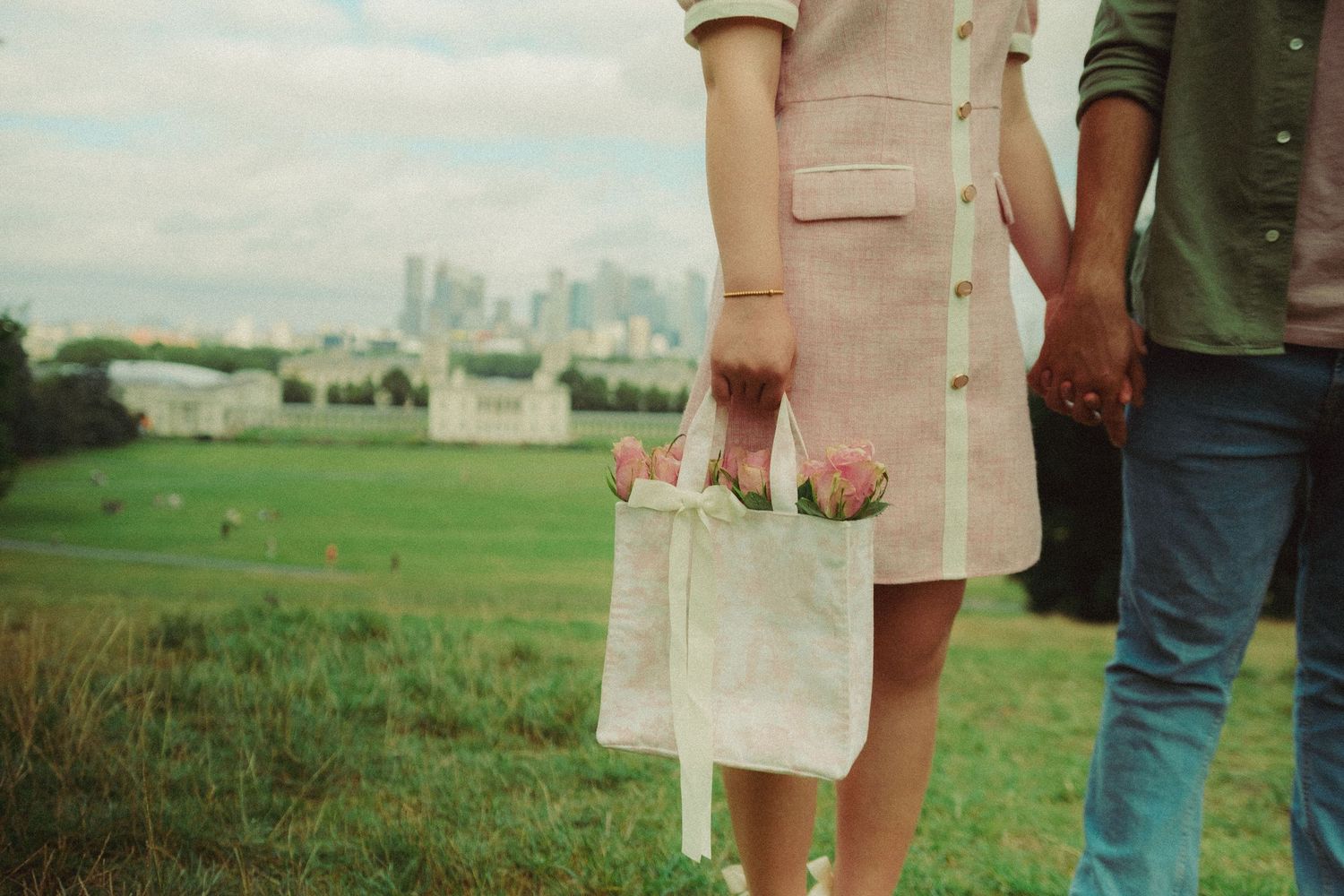 Candid pre-wedding mid close up photograph of a couple walking hand in hand through a green park with the city skyline in the background.