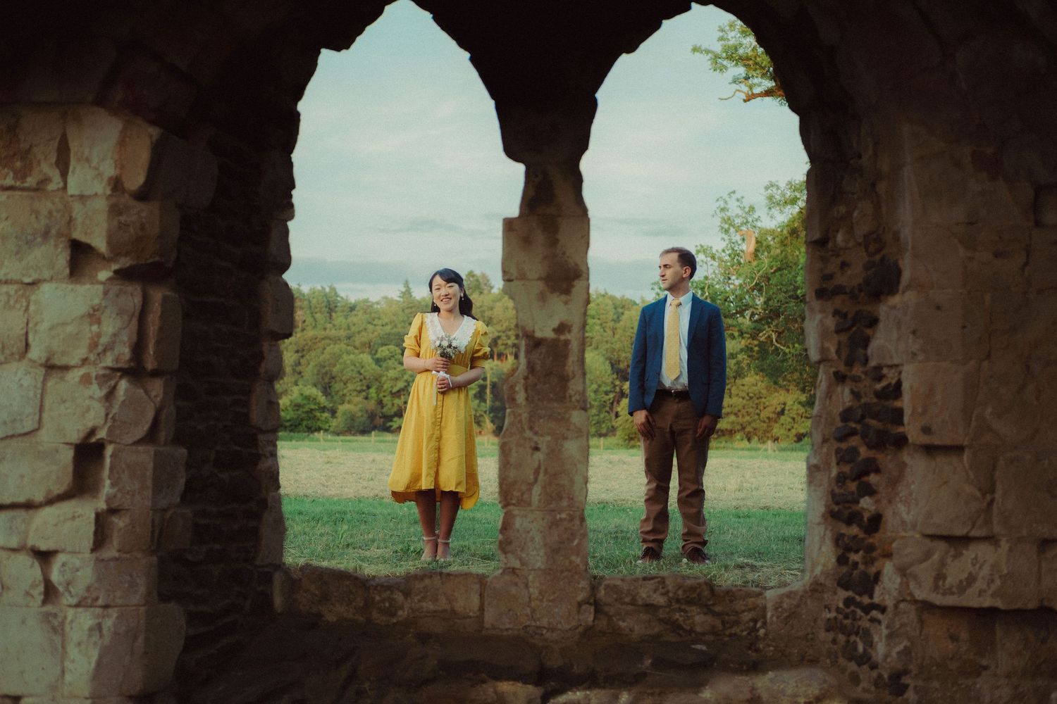 Cinematic pre-wedding photograph of a couple standing together through the archway of a historic stone building.
