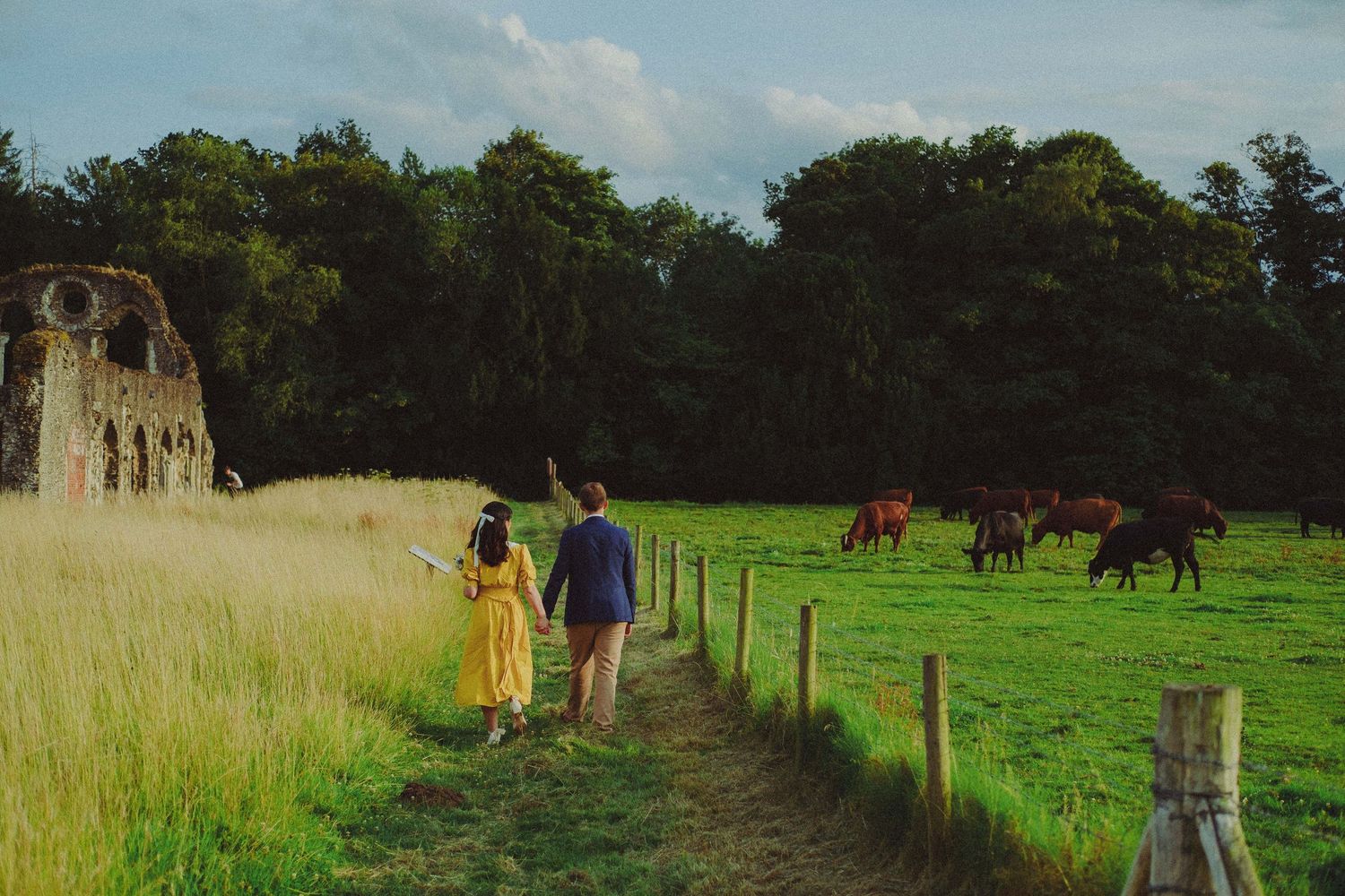 Cinematic pre-wedding photograph of a couple walking together through a countryside field beside a historic building.