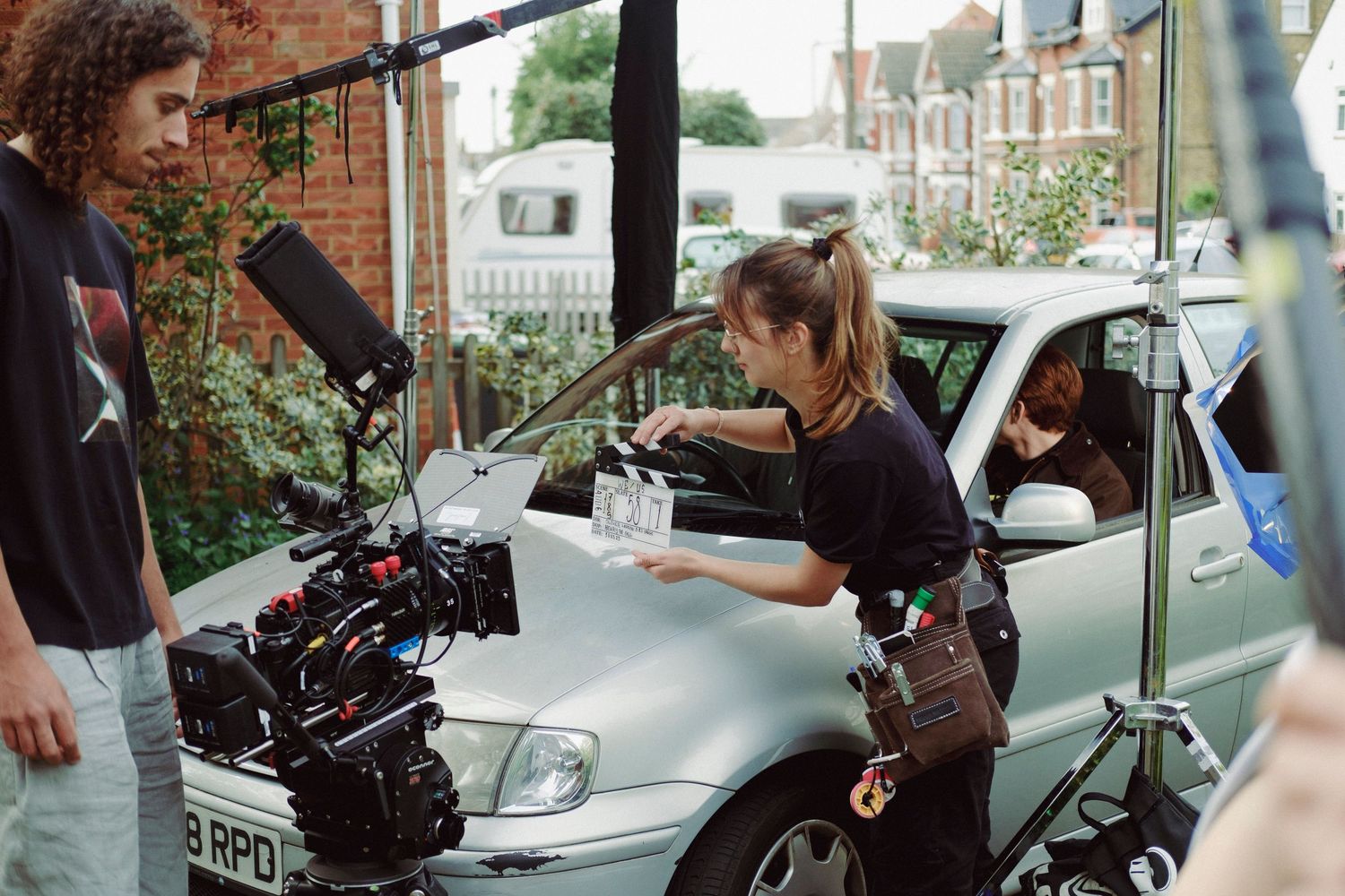 Behind-the-scenes photograph of a film crew shooting a scene with a professional cinema camera beside a car on location.