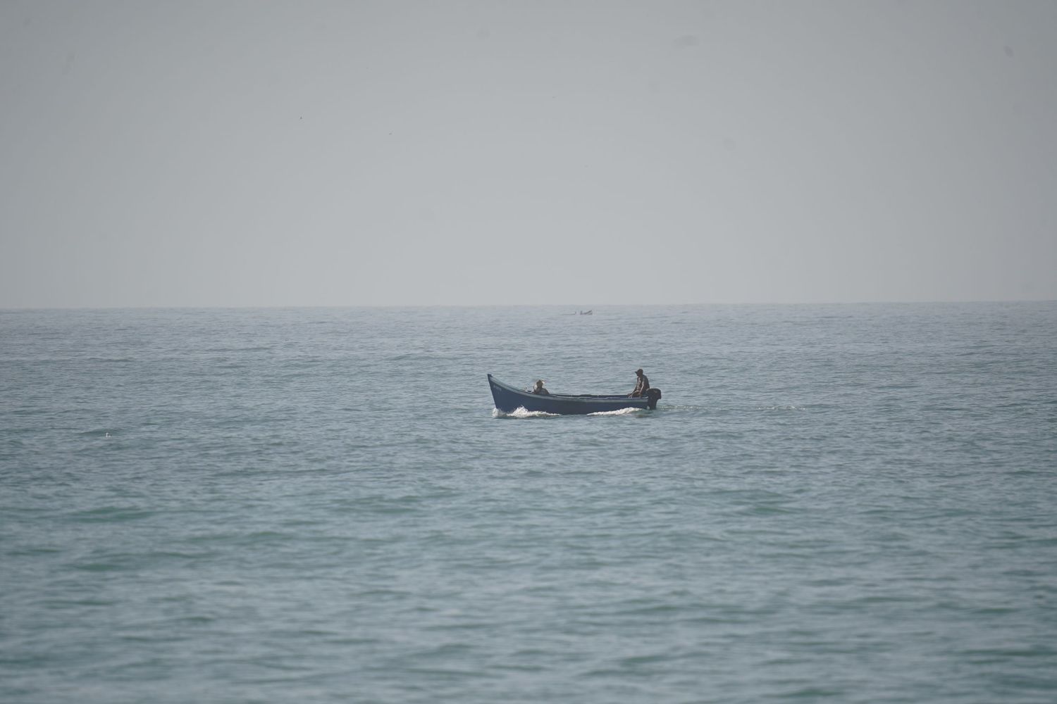 Lone fisherman in a small wooden boat on calm open ocean under hazy grey sky.