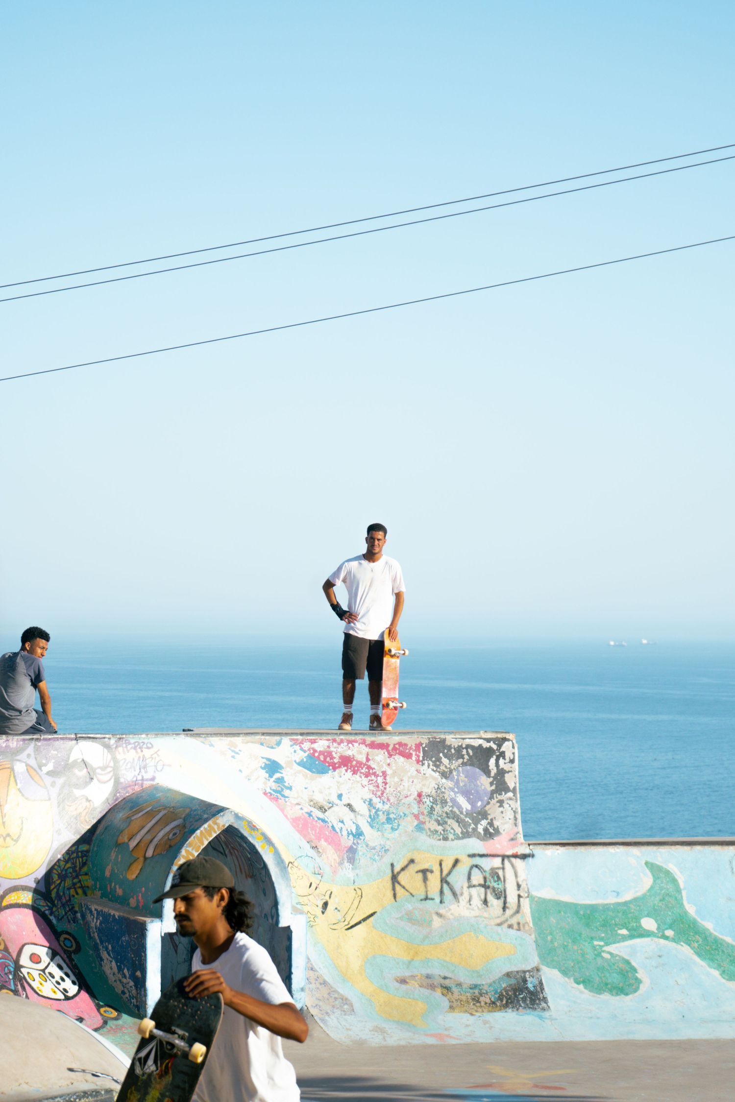 Skateboarders at graffiti-covered oceanside skate park, one standing on ramp edge holding longboard above blue sea.