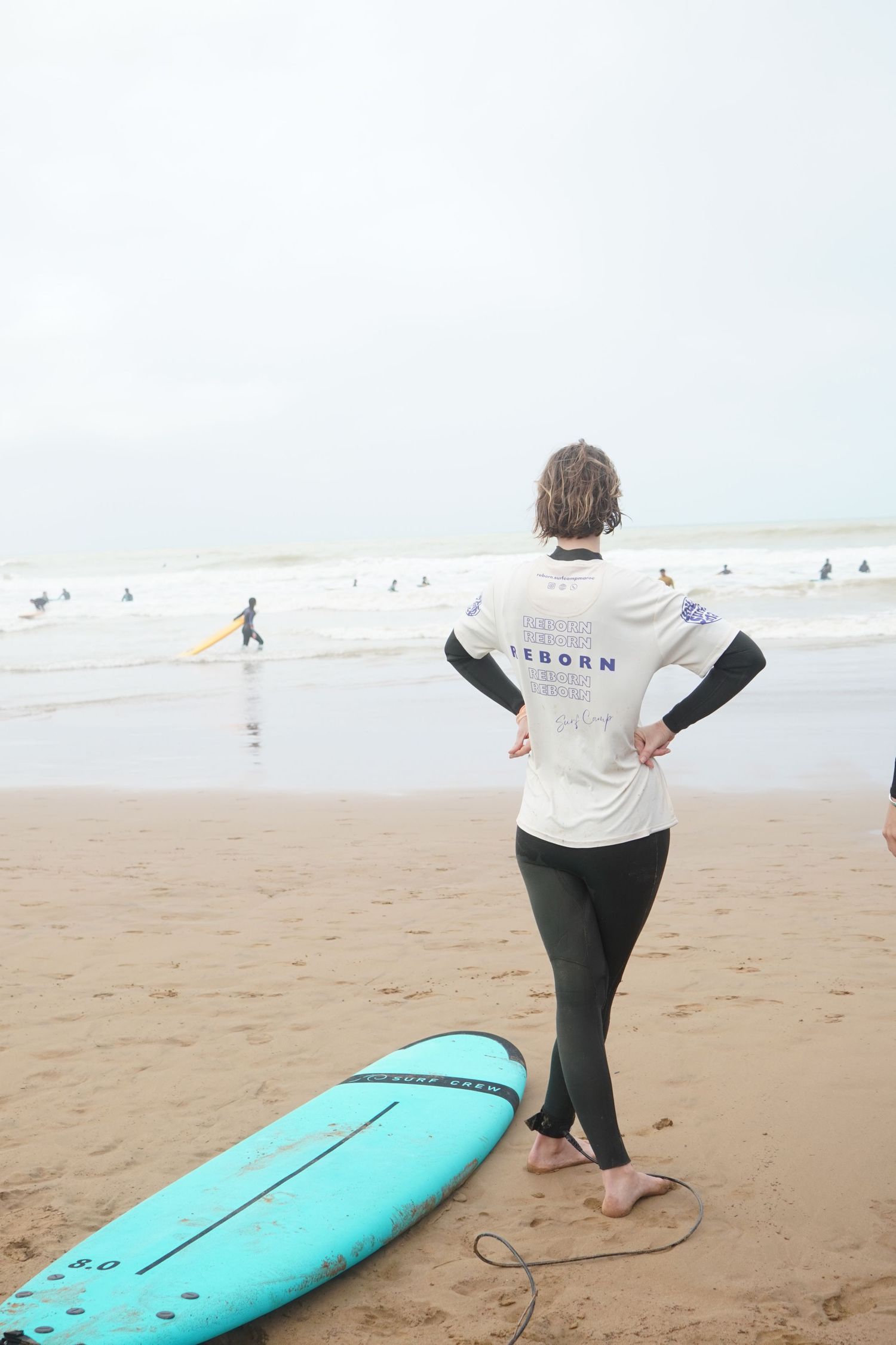 Surfer in white Reborn jersey stands on sandy beach beside turquoise surfboard, watching waves on overcast day.