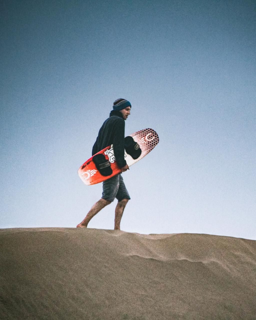 A man carrying a red surfboard walks barefoot across sandy dunes under a clear blue sky.