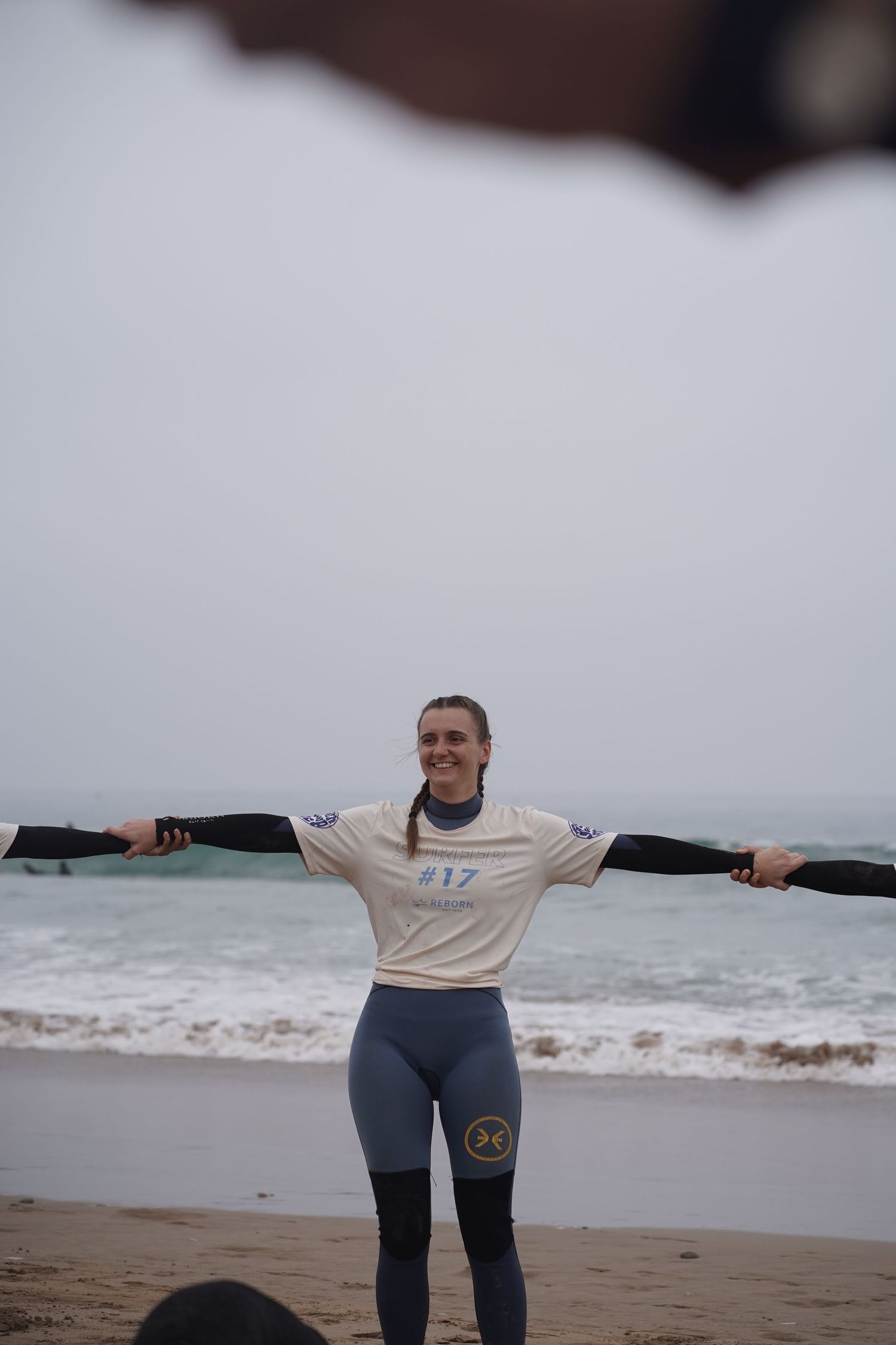 Woman in #17 jersey holds oars wide on a foggy beach, smiling confidently near ocean waves.