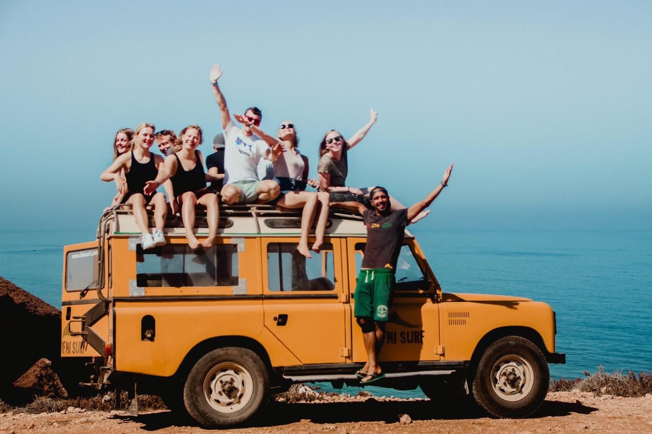 Group of friends sitting on top of a yellow Land Rover on a coastal cliff with blue ocean views.