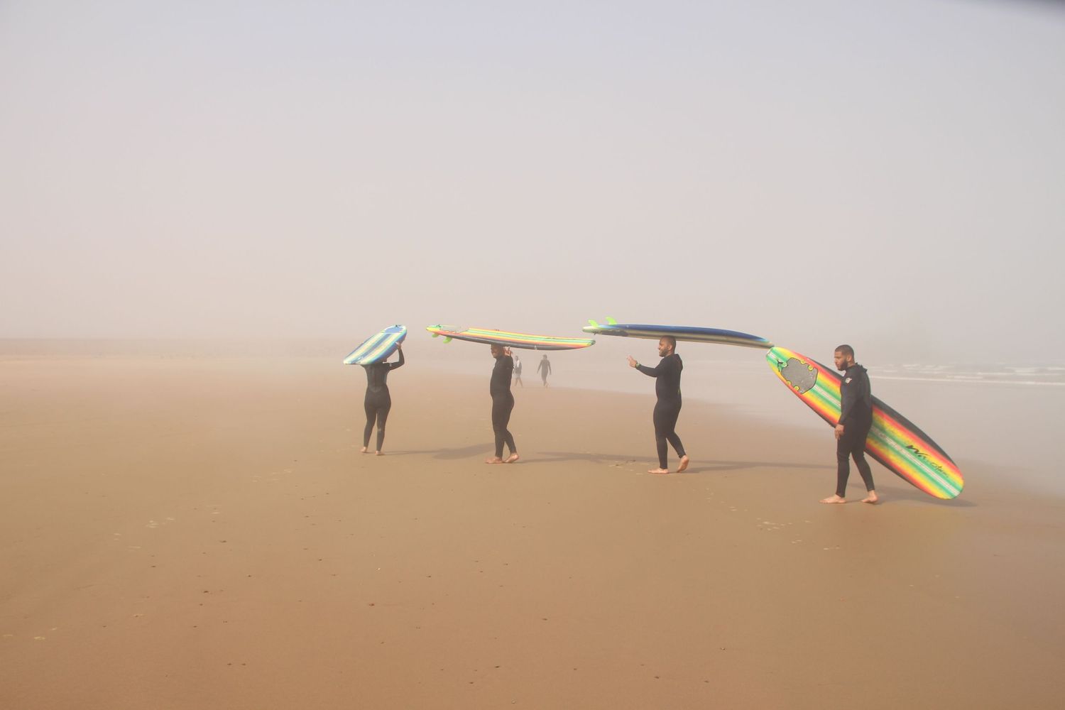 Four surfers in wetsuits carry colorful surfboards on a misty sandy beach.