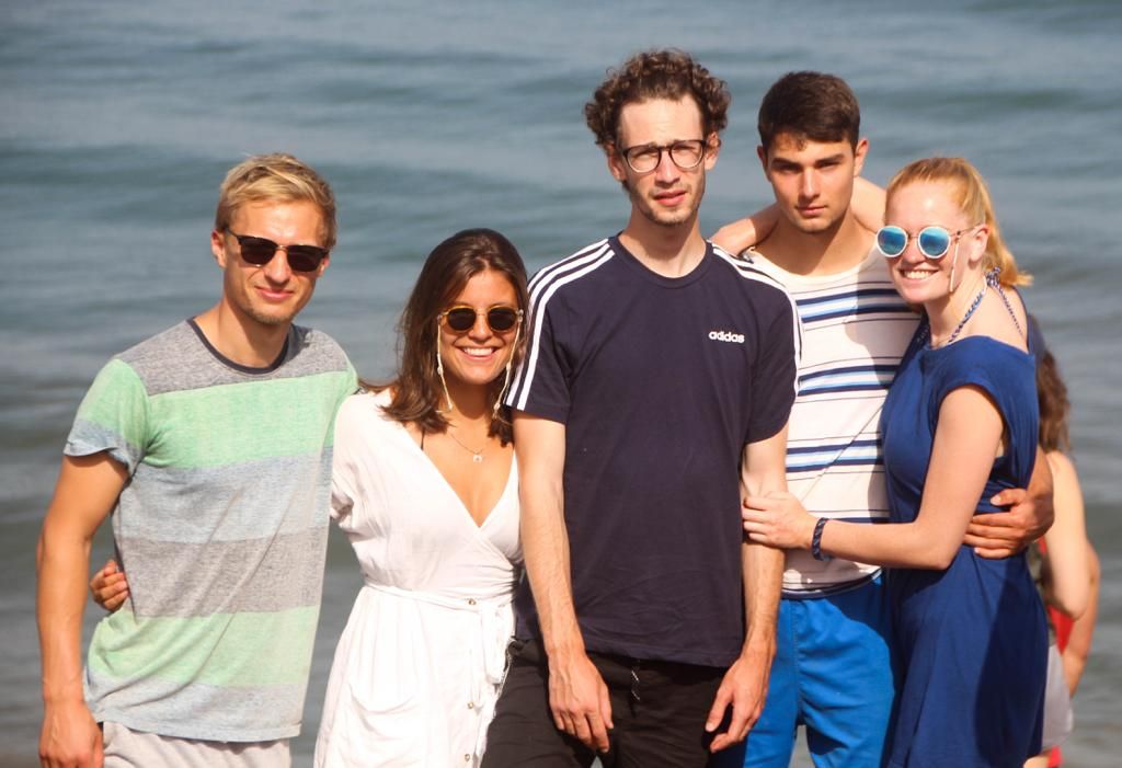 Five young adults posing together on a sunny beach with ocean waves in the background.