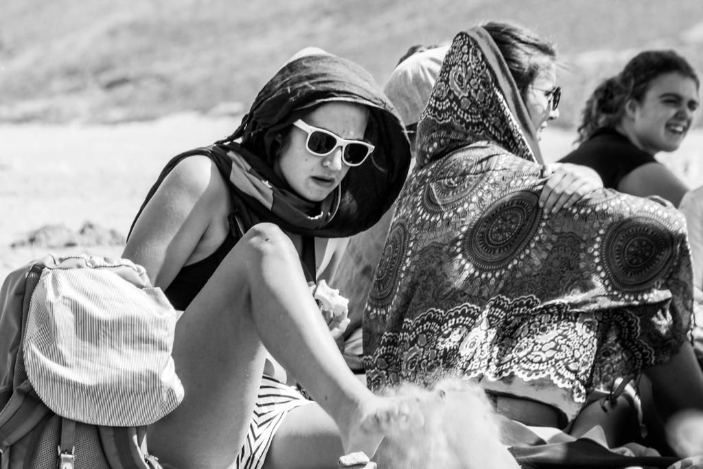 Black and white photo of a woman wearing sunglasses and a head scarf sitting on a beach with others.