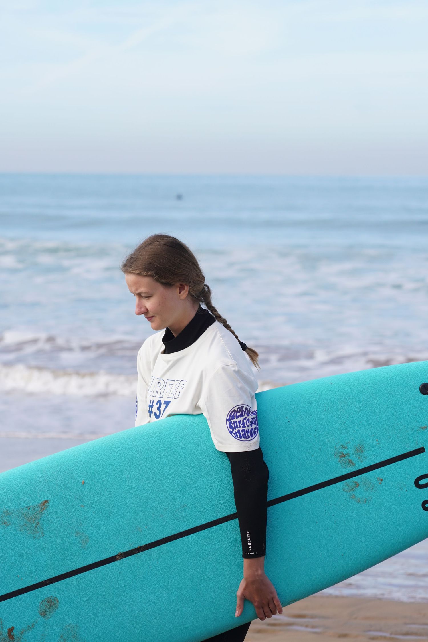Young female surfer wearing #37 competition jersey carries a turquoise longboard along a sandy beach.
