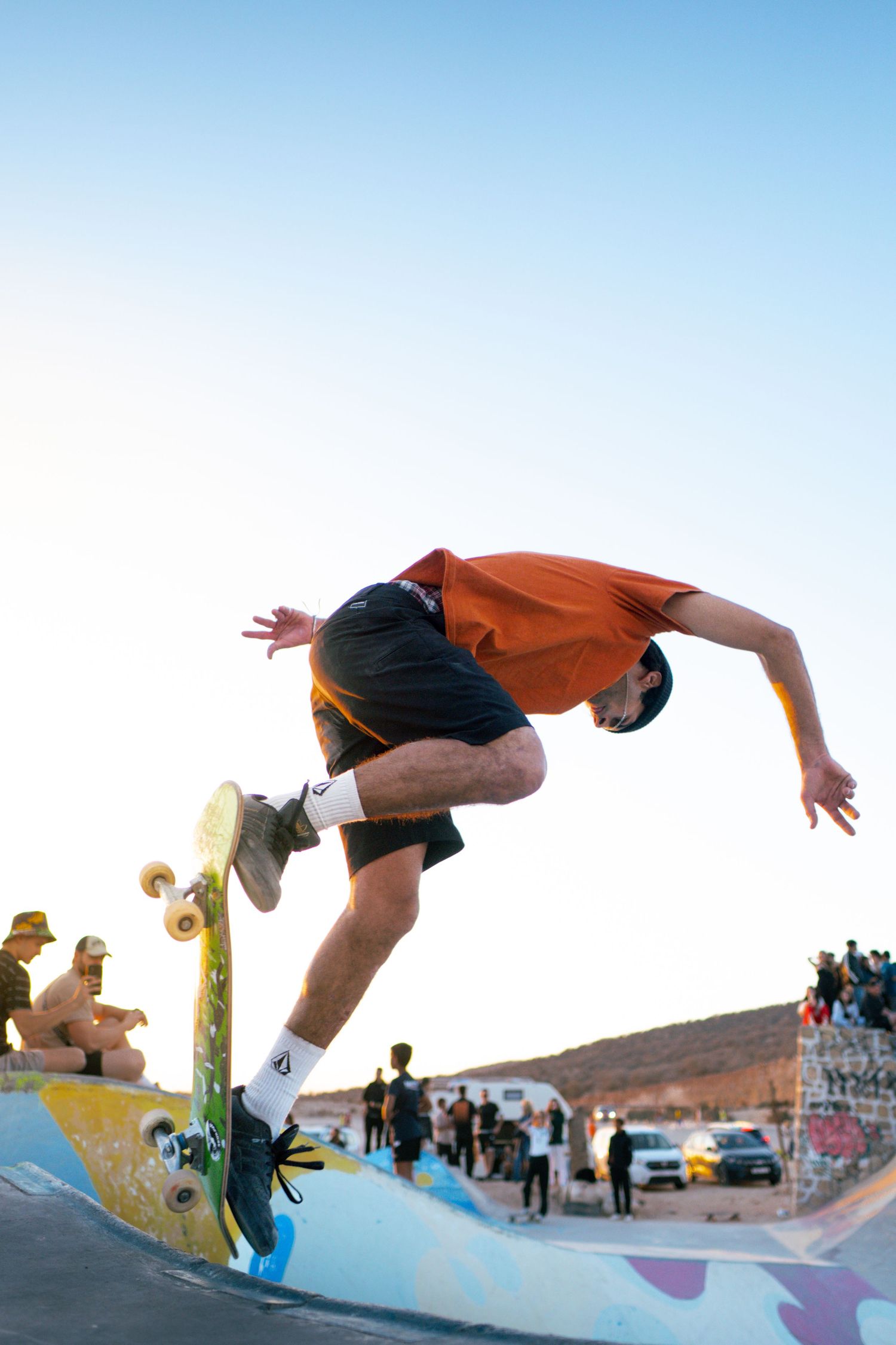 Skateboarder in orange shirt executes aerial trick on colorful ramp at outdoor skate park during golden hour.