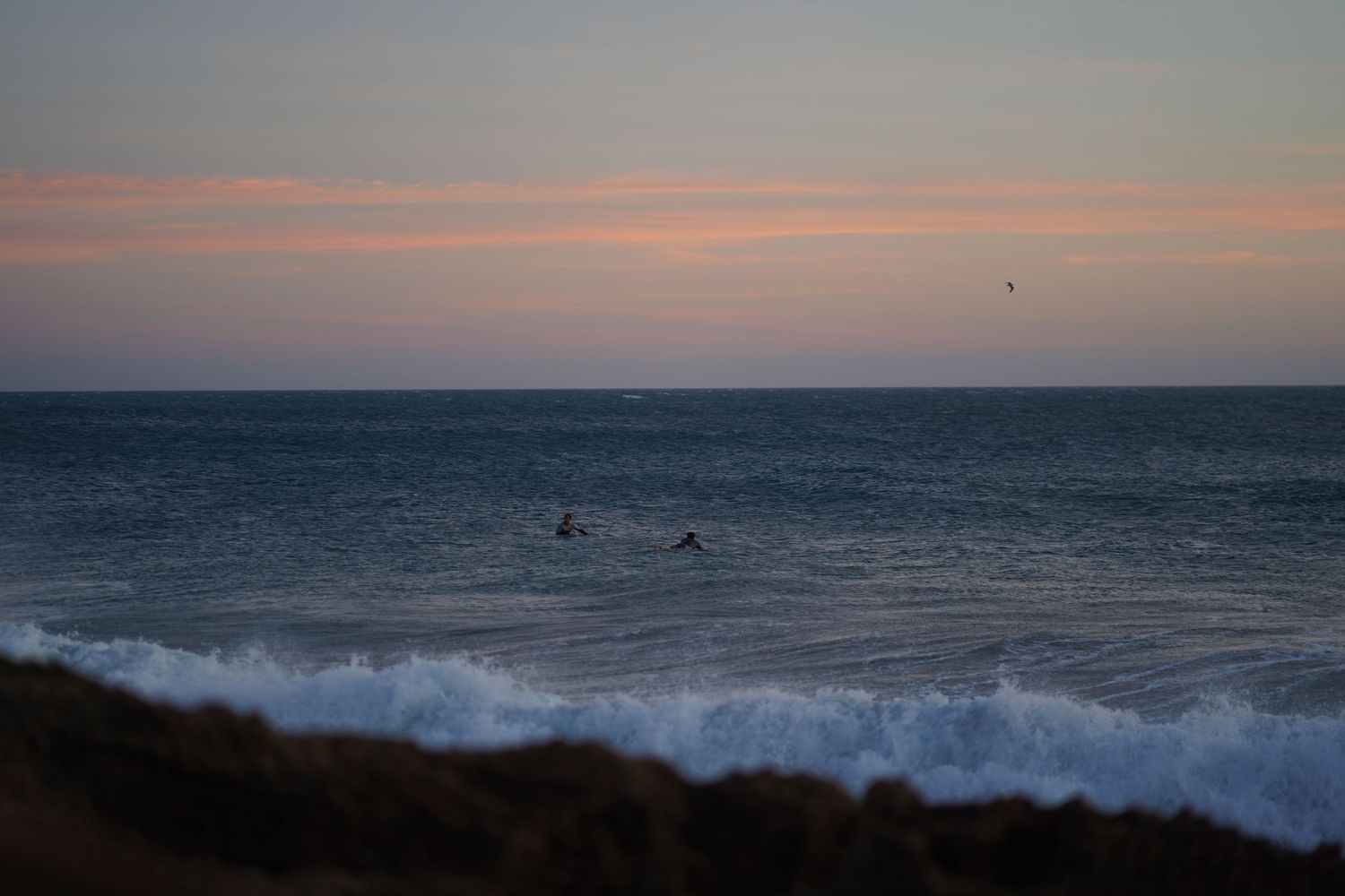 Surfers ride waves at sunset with colorful pink and orange sky over the ocean.