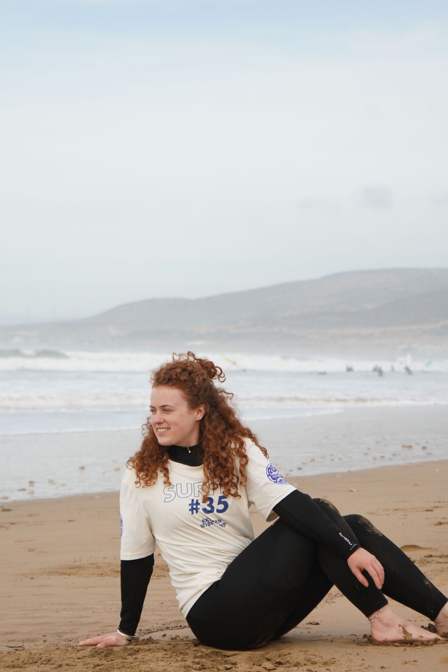 Young woman with curly red hair sits smiling on sandy beach wearing white rash guard #35 and black wetsuit pants.