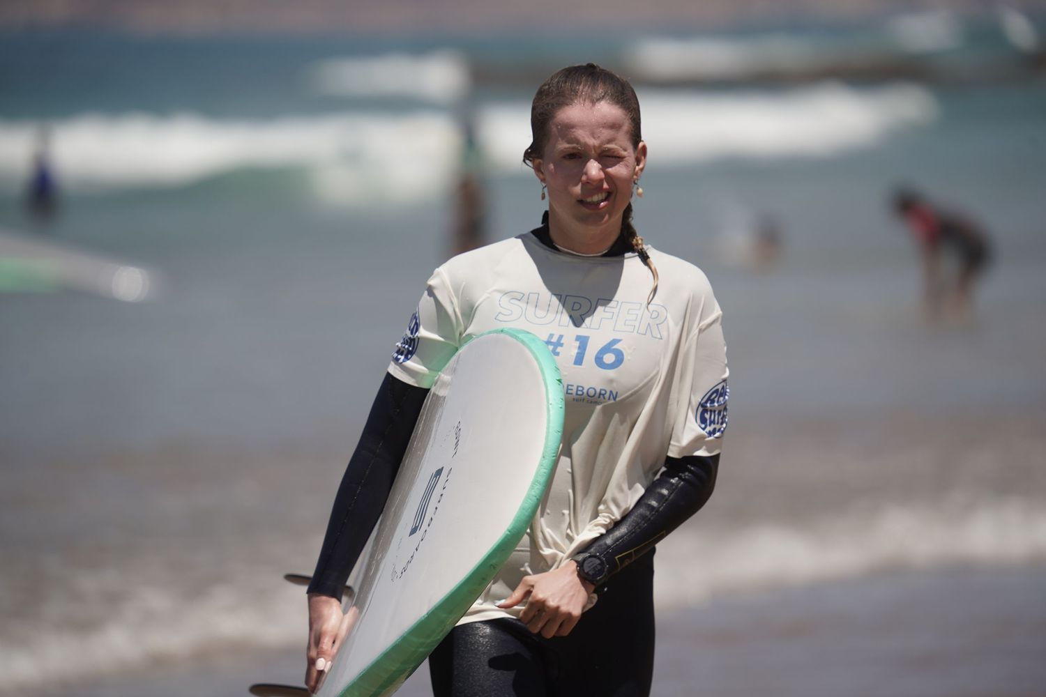 Young female surfer in wetsuit carrying a white surfboard on a sunny beach near ocean waves.
