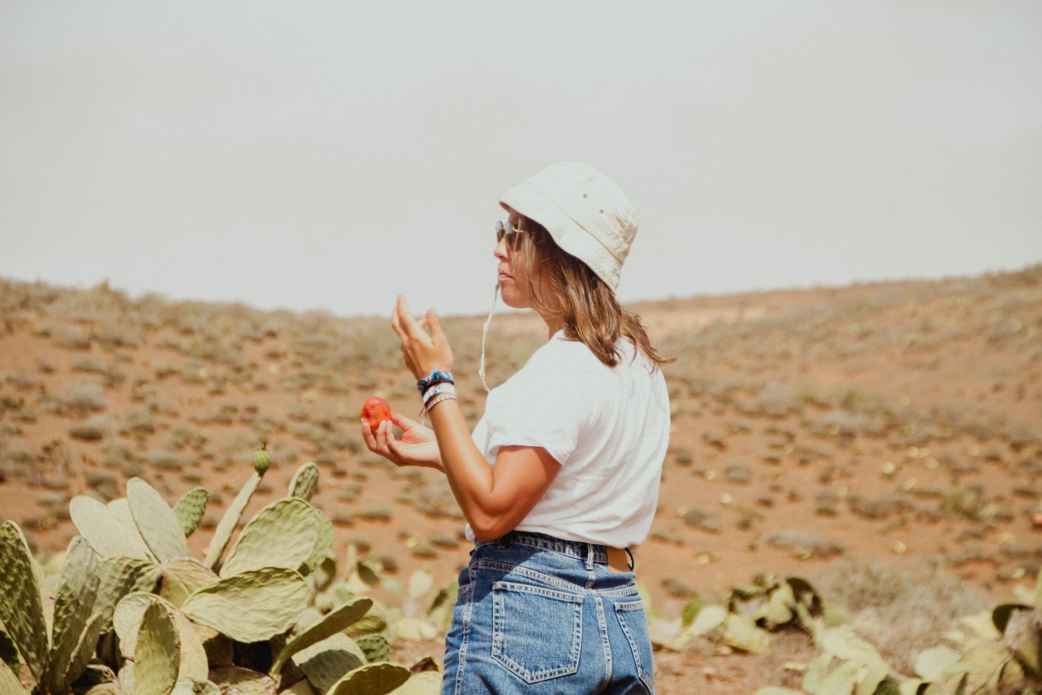 Woman in white hat and jeans holding cactus fruit in a desert landscape with cacti.