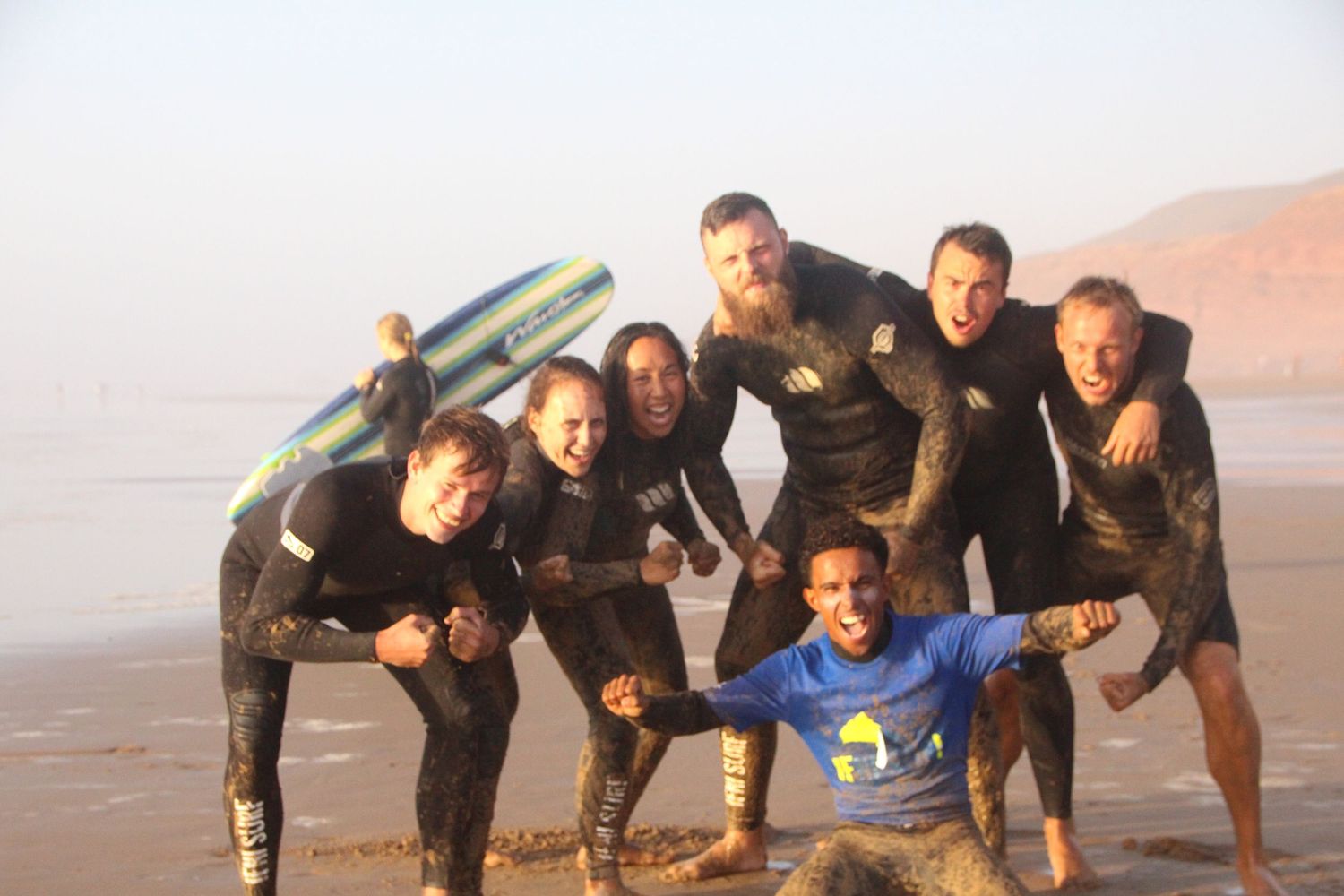 Group of surfers in wetsuits posing together on a foggy beach, smiling and holding a surfboard.