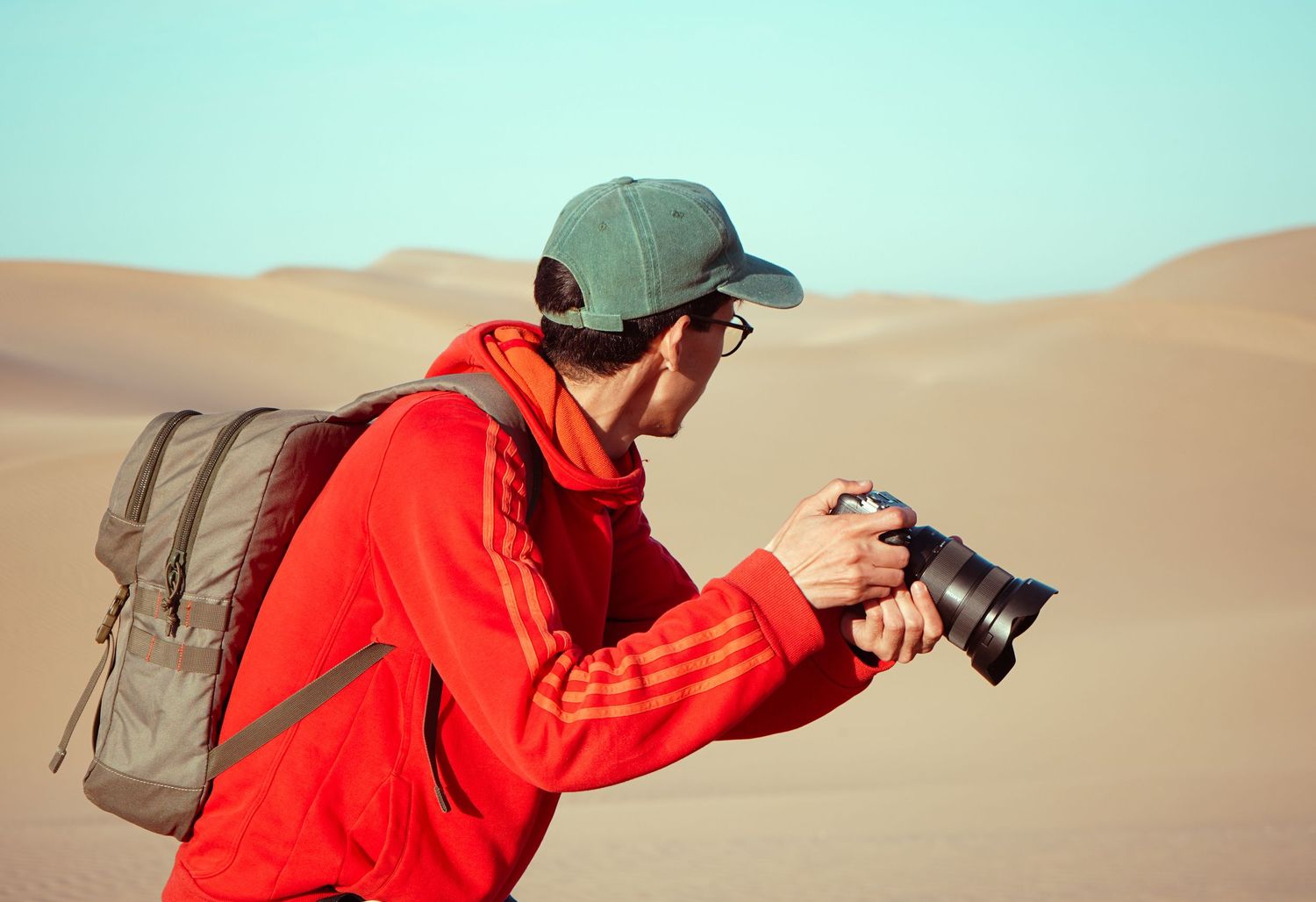 A photographer in a red Adidas hoodie and cap reviews shots on his camera while exploring sandy desert dunes.