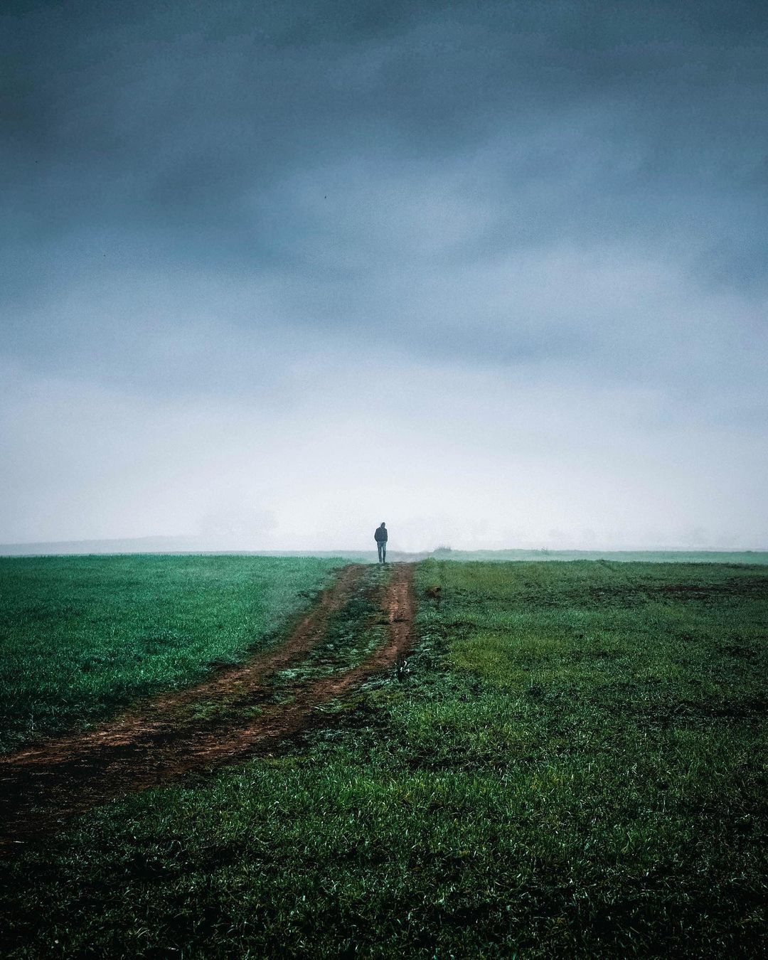 A solitary figure stands on a dirt path through green fields under a dramatic stormy sky.