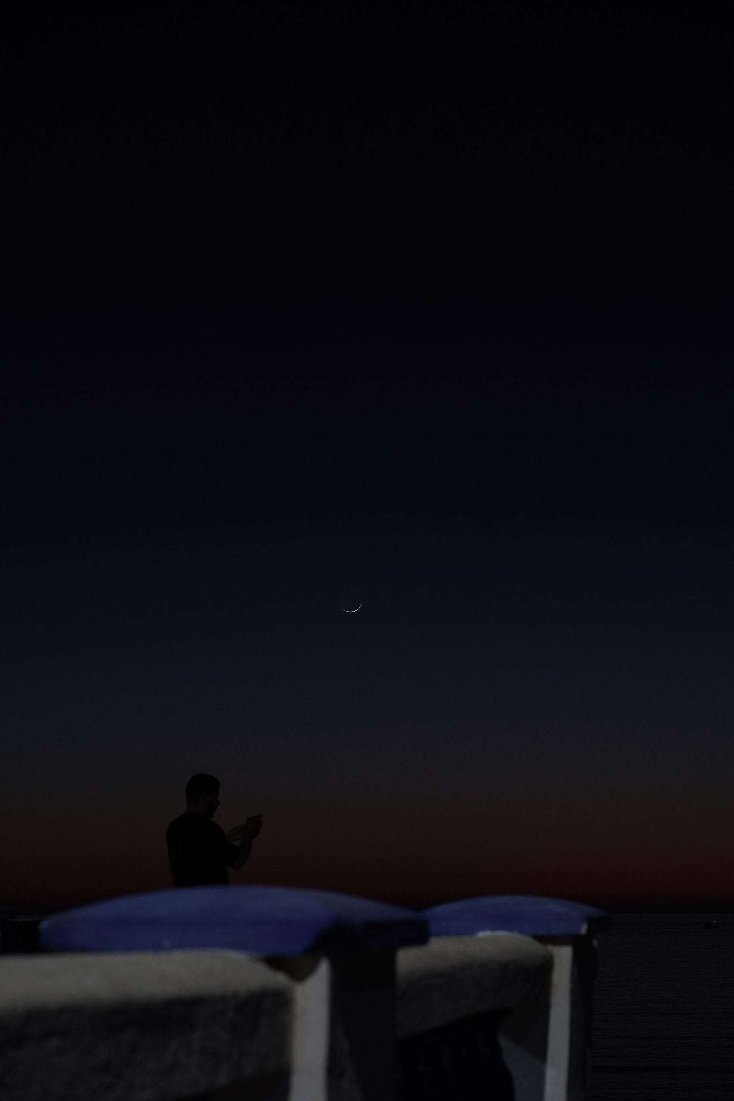 Silhouette of person sitting at night with crescent moon visible in dark twilight sky.