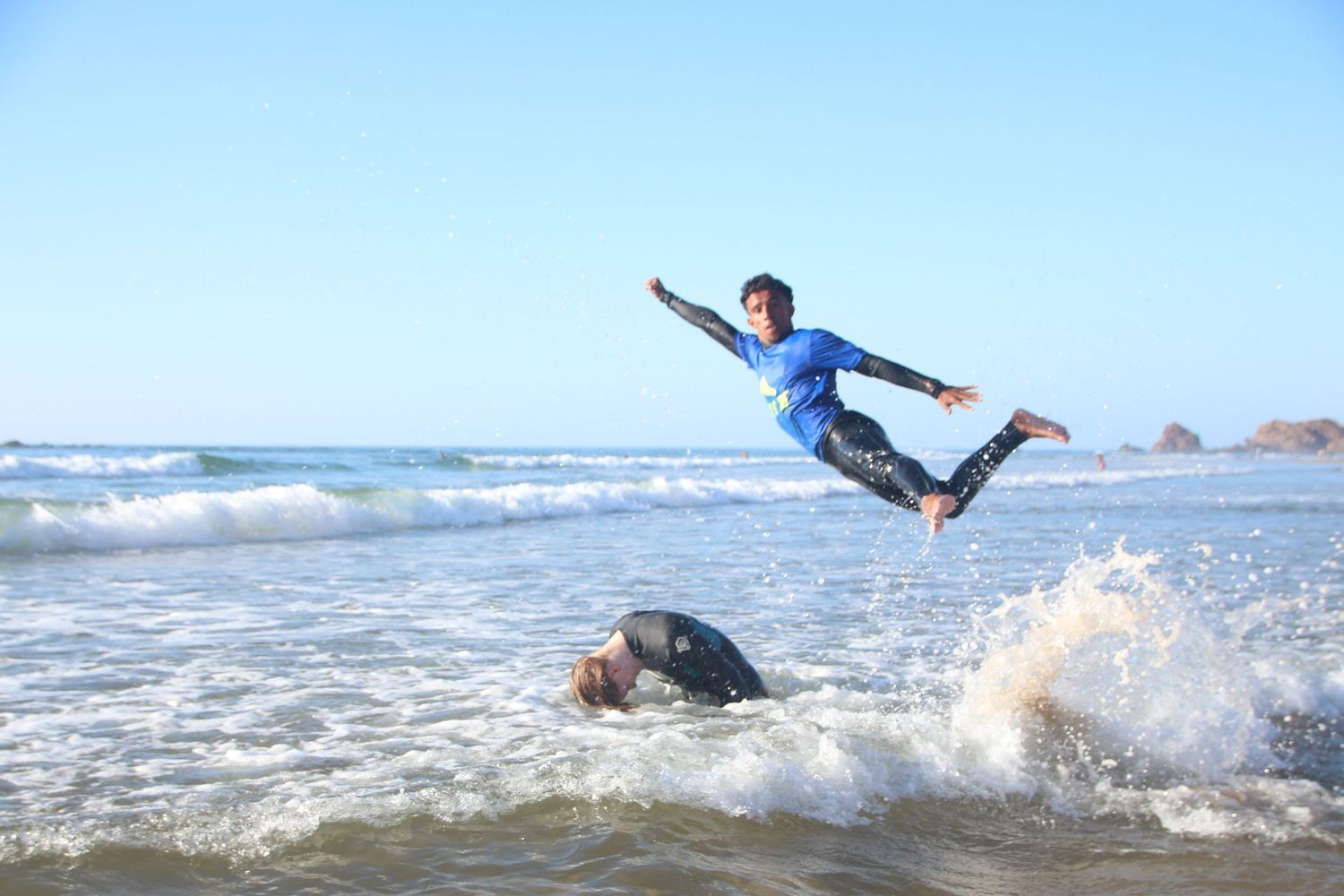 A surfer leaps into the air above crashing waves while another person lies in the shallow ocean water on a sunny day.