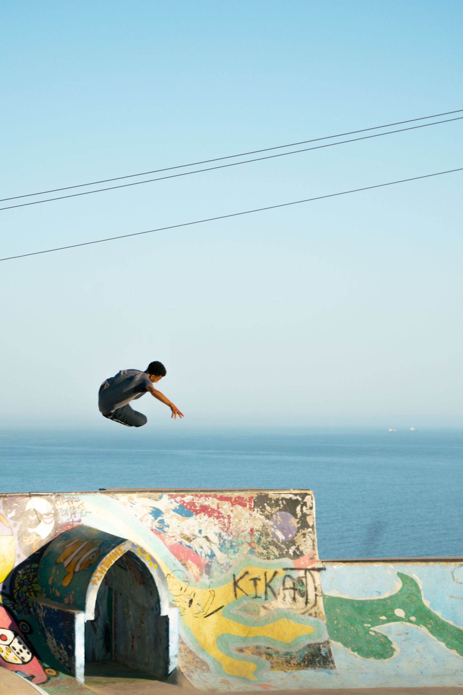 Skateboarder launches high above graffiti-covered ramp at oceanside skate park with ocean visible in background.