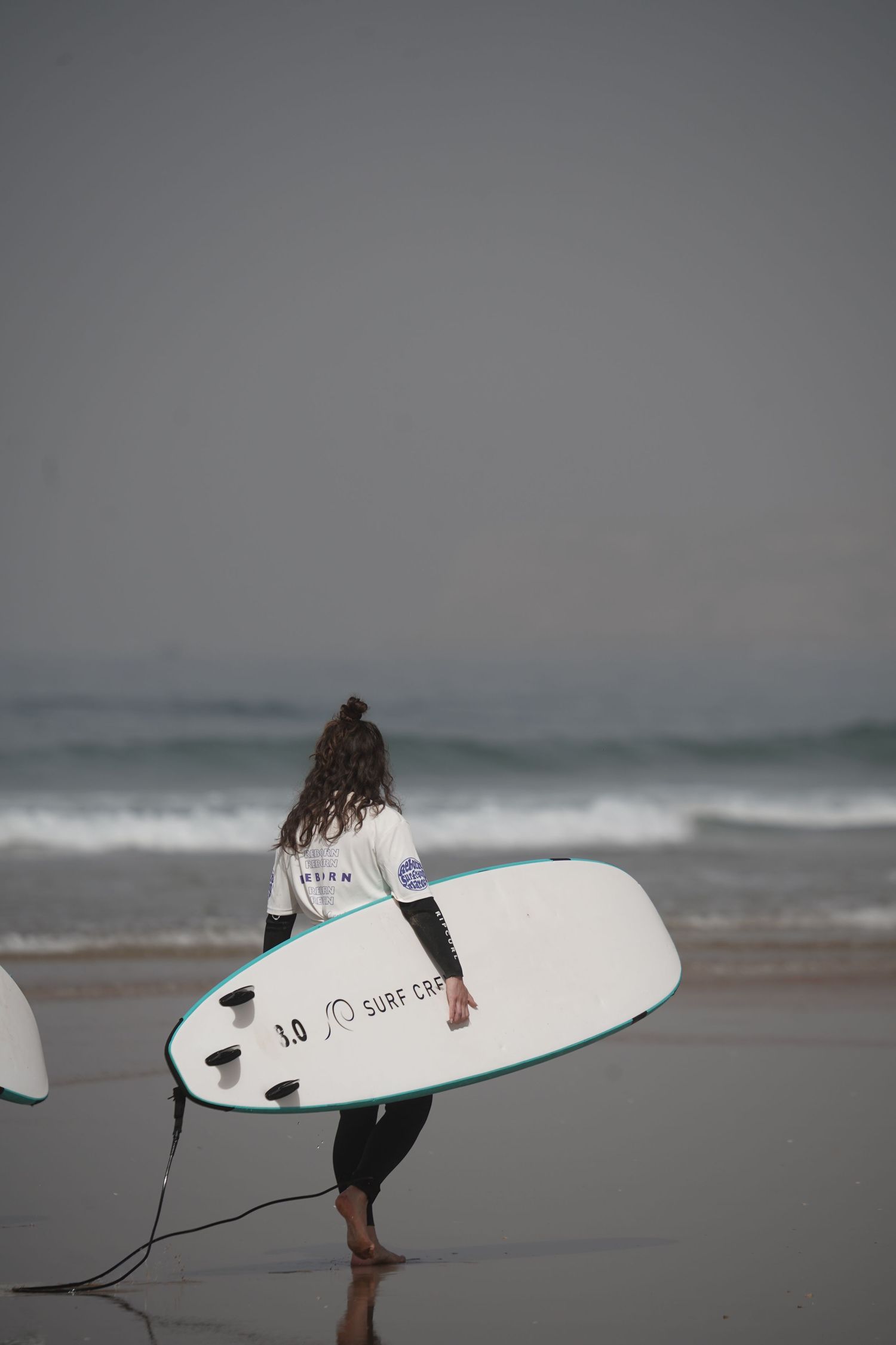 A young woman with curly hair carries a white surfboard toward the ocean on a misty beach wearing a wetsuit.