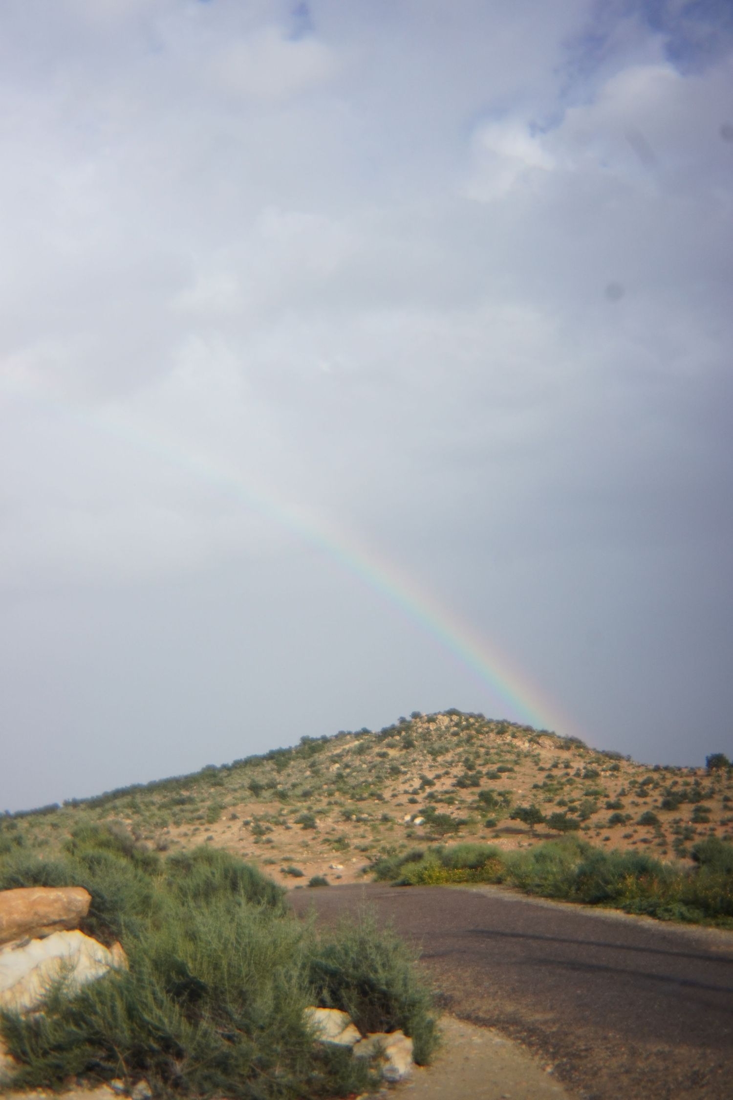 Colorful rainbow arching over a rocky desert hill with sparse vegetation and winding road.