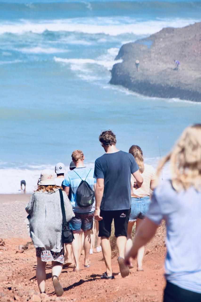 Group of tourists walking down a red sandy path toward a beautiful blue ocean beach on a sunny day.