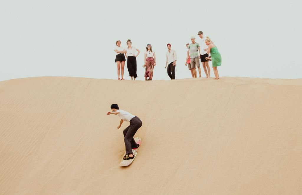 A person sandboarding down a large sand dune while a group of spectators watches from the top.