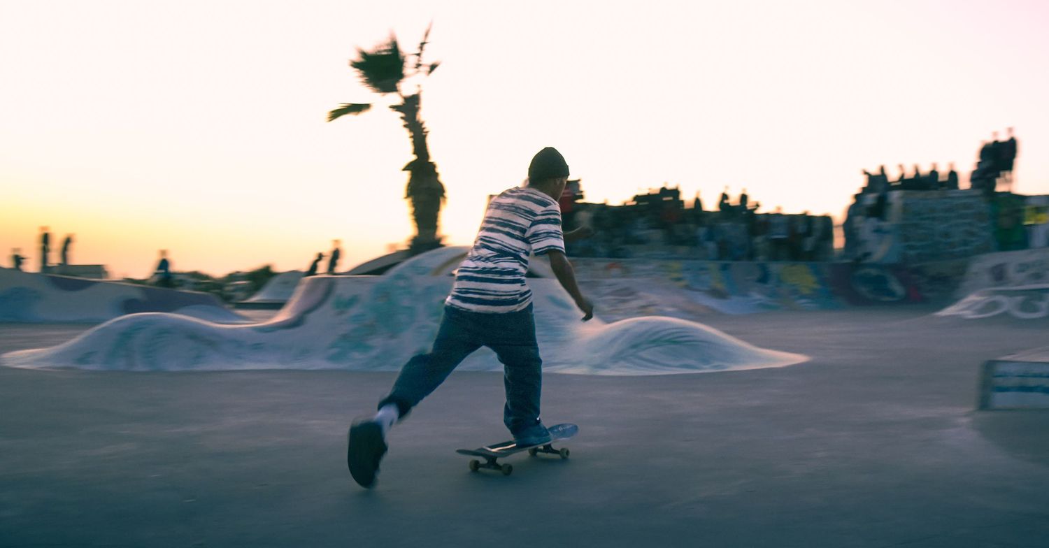 Skateboarder riding through an outdoor skate park at golden hour with a palm tree silhouette in background.