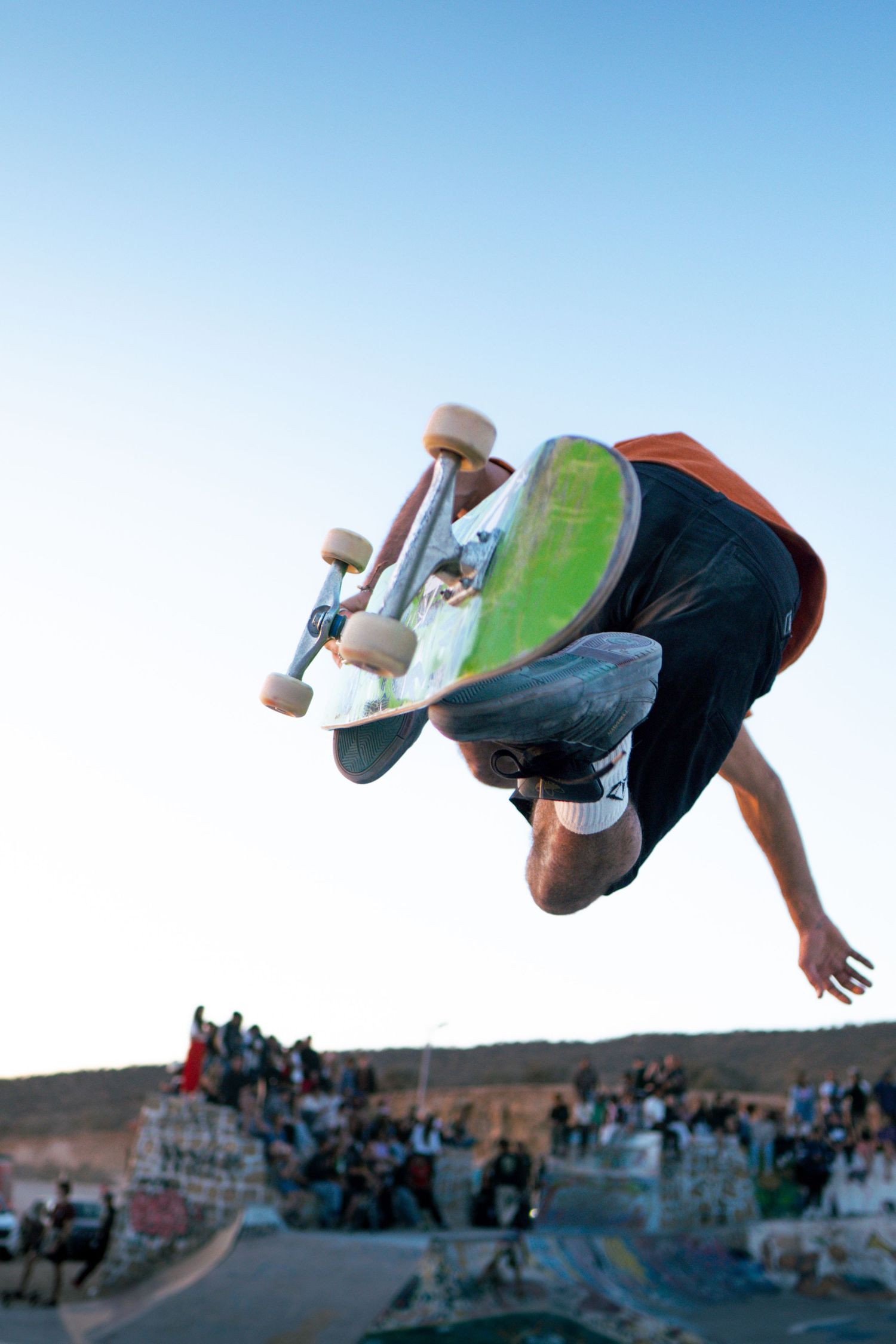 Skateboarder performs inverted aerial trick high above outdoor skate park with crowd visible in background.