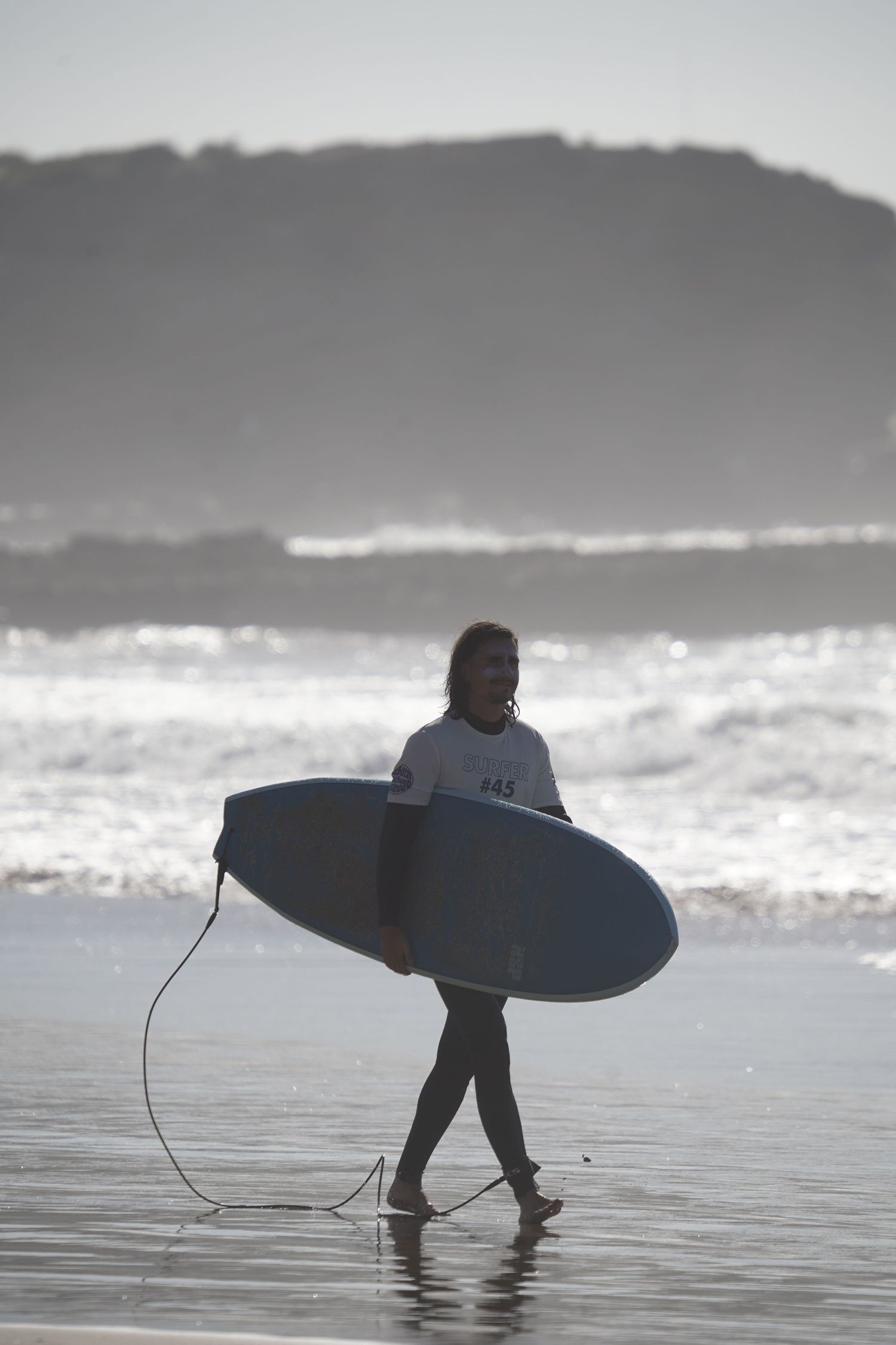 Surfer carrying a blue surfboard walks along a misty beach with ocean waves and a mountain in the background.