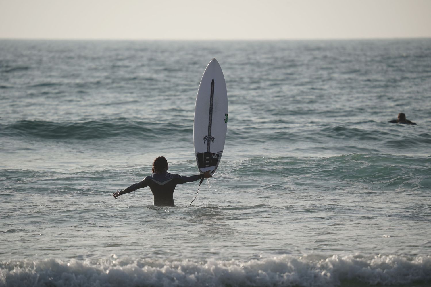 Surfer holding a white surfboard upright while wading into ocean waves at dusk.