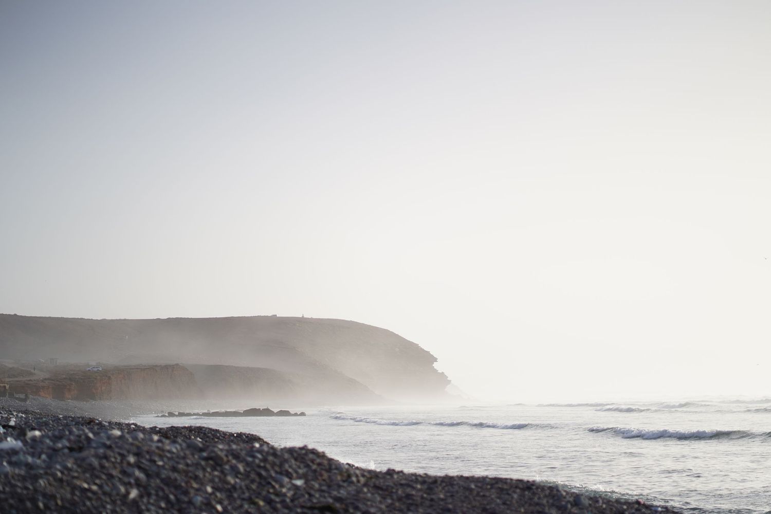Misty coastal cliffs rise above rocky shores with gentle ocean waves under a hazy sky.