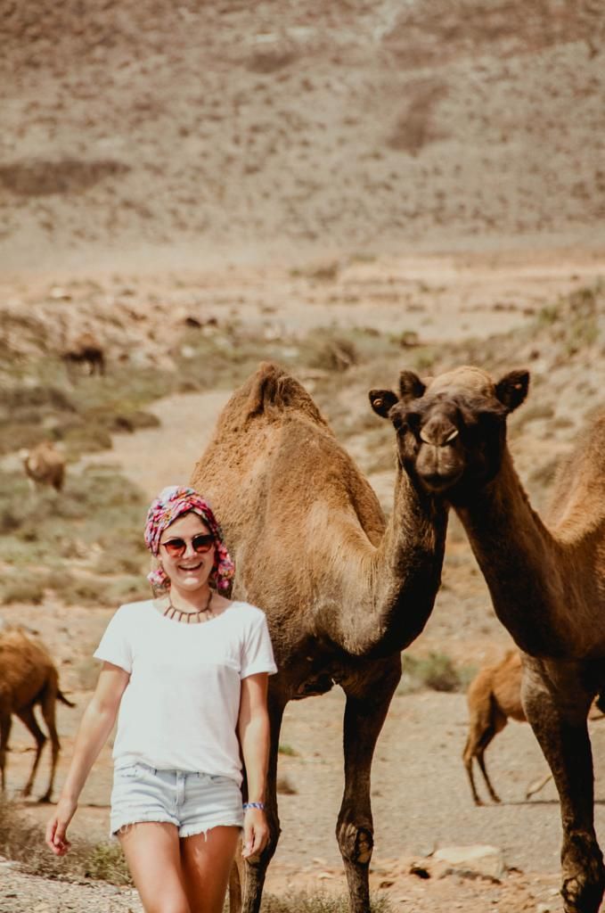 A smiling woman in white tee and denim shorts poses beside a camel in a desert landscape.