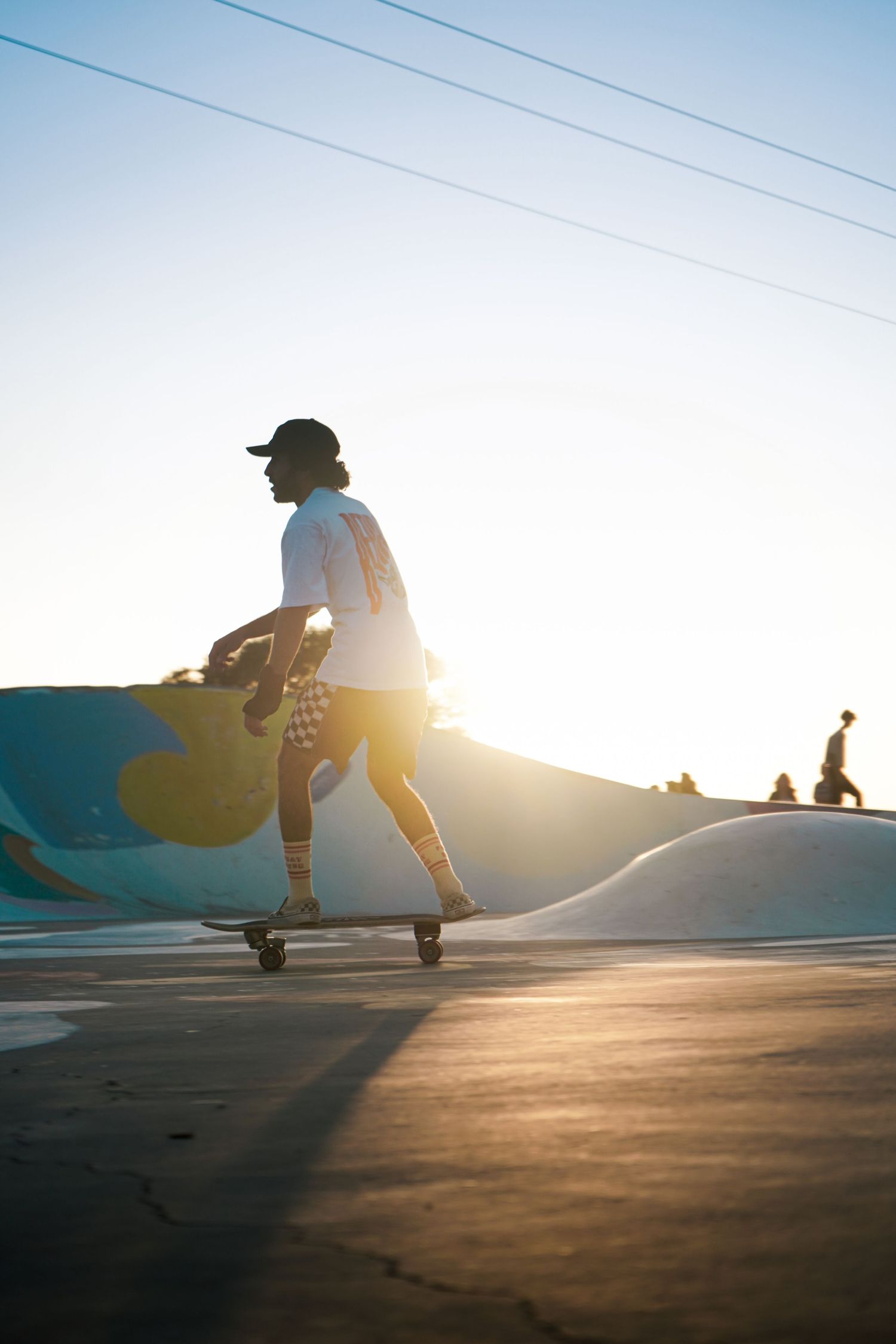 Silhouetted skateboarder cruising past a colorful mural wall at a sun-drenched outdoor skate park.