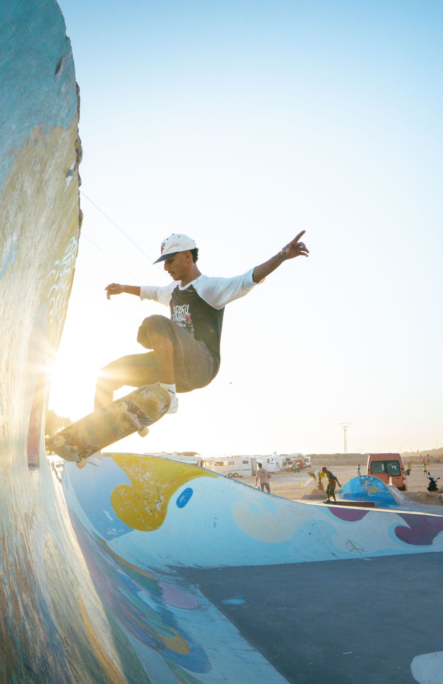 Young skateboarder performing trick on colorful painted ramp wall at sunset skatepark with bright backlight.