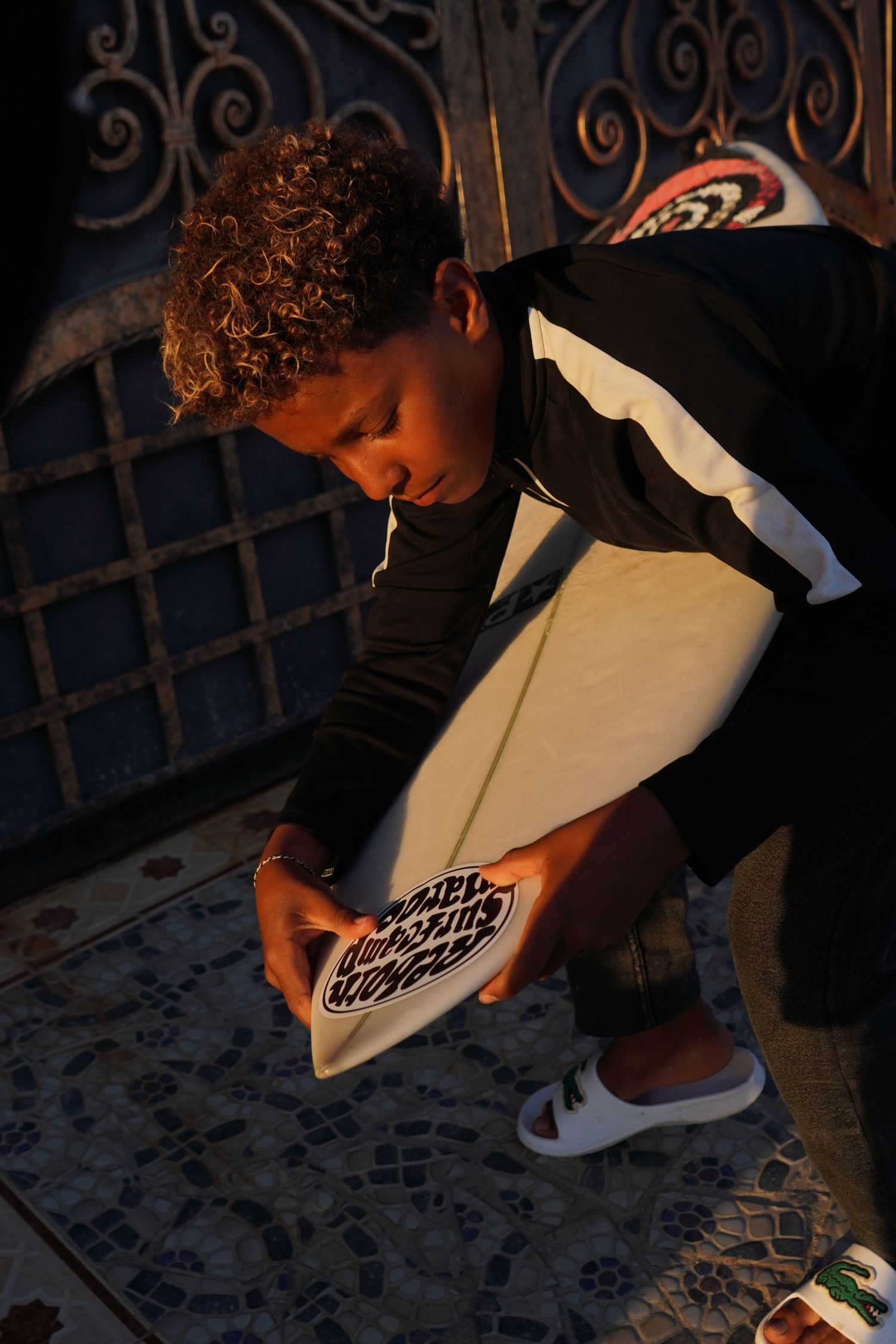 Young surfer with curly hair crouching to examine a white surfboard near an ornate metal gate.