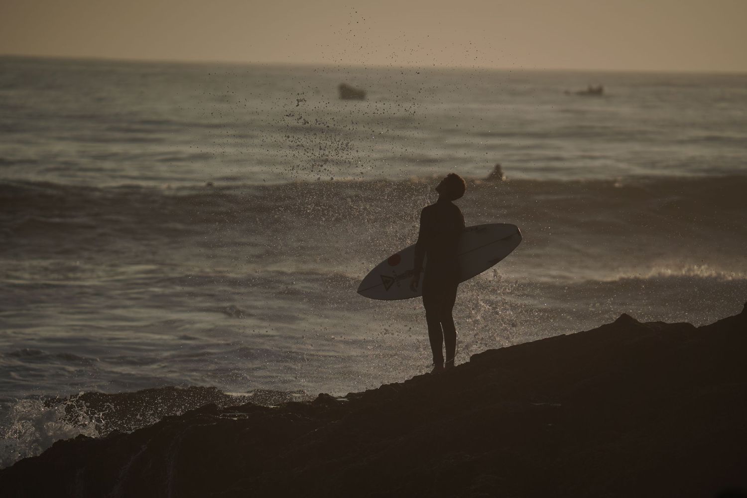 Silhouette of a surfer holding a board on rocky shore at dusk with ocean waves in the background.