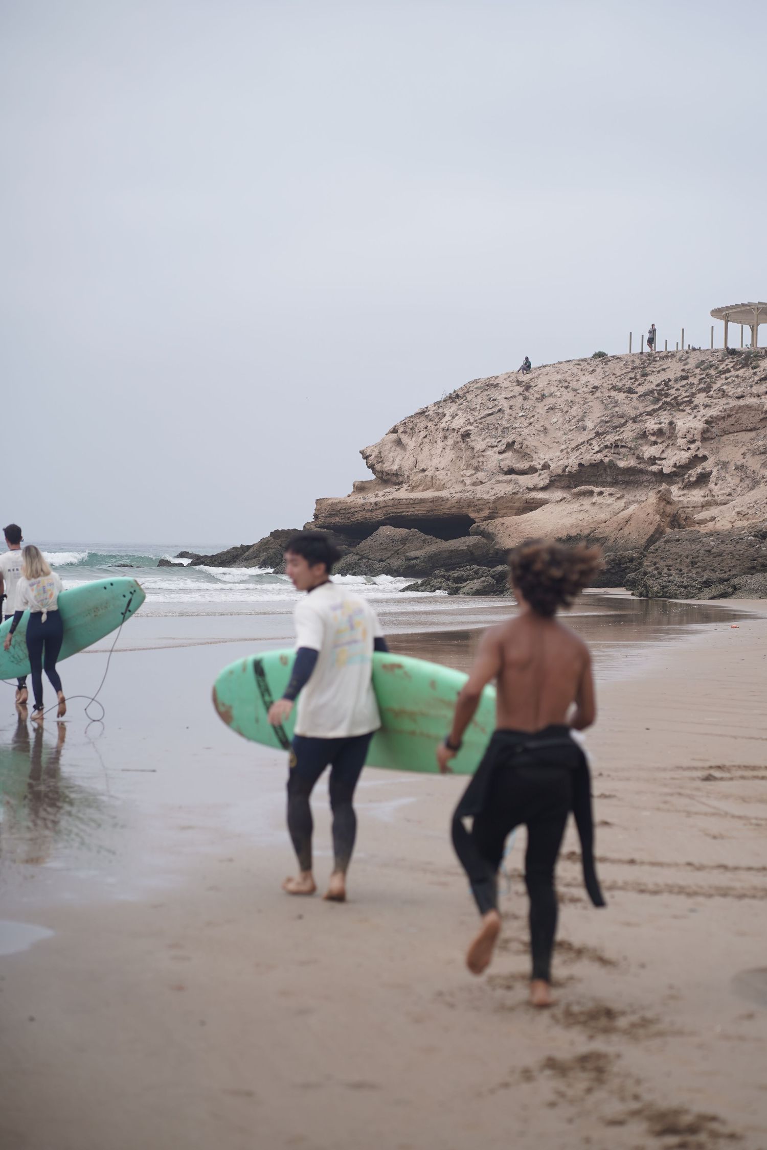 Surfers carrying green boards walk along a sandy beach toward rocky cliffs on an overcast day.