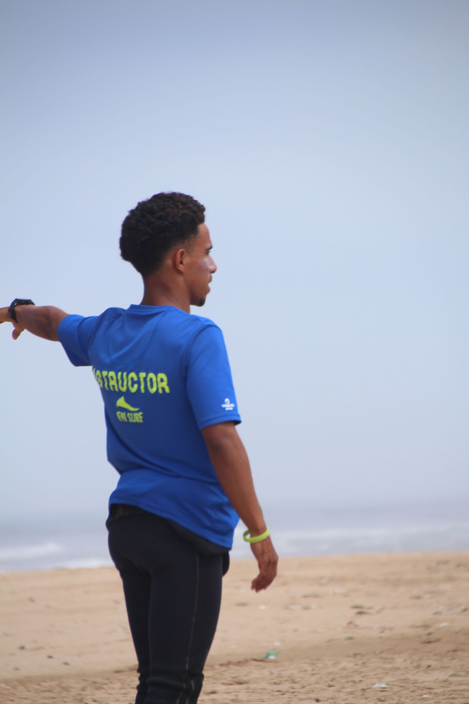 A surf instructor in a blue shirt gestures on a sandy beach with a misty ocean backdrop.