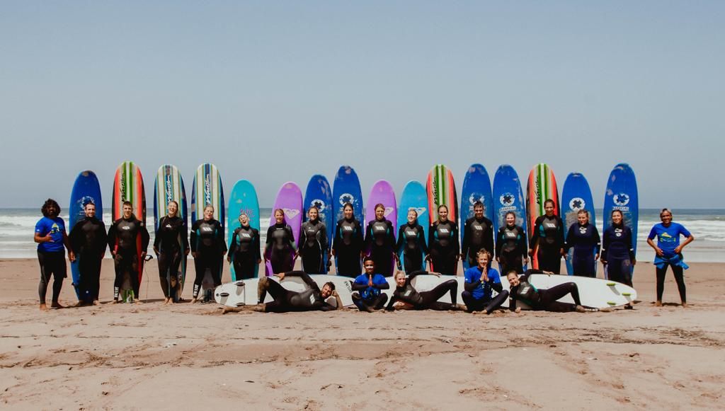 Large group of surfers posing with colorful surfboards on a sandy beach under a clear blue sky.