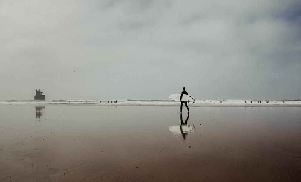 A surfer walks along a misty reflective beach with calm waves and overcast skies in the background.