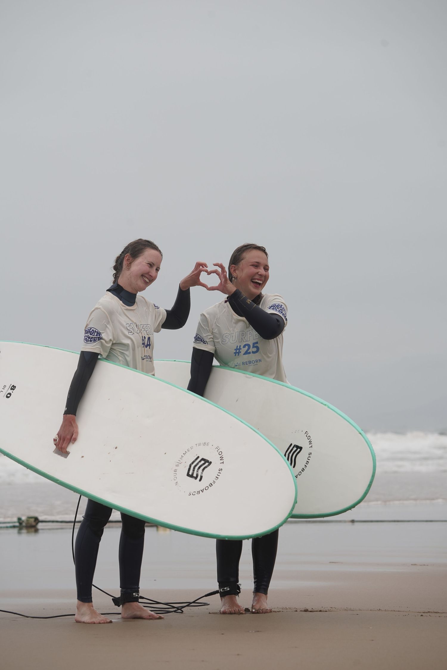 Two smiling female surfers in wetsuits holding white boards on a beach, one making a heart shape with her hands.